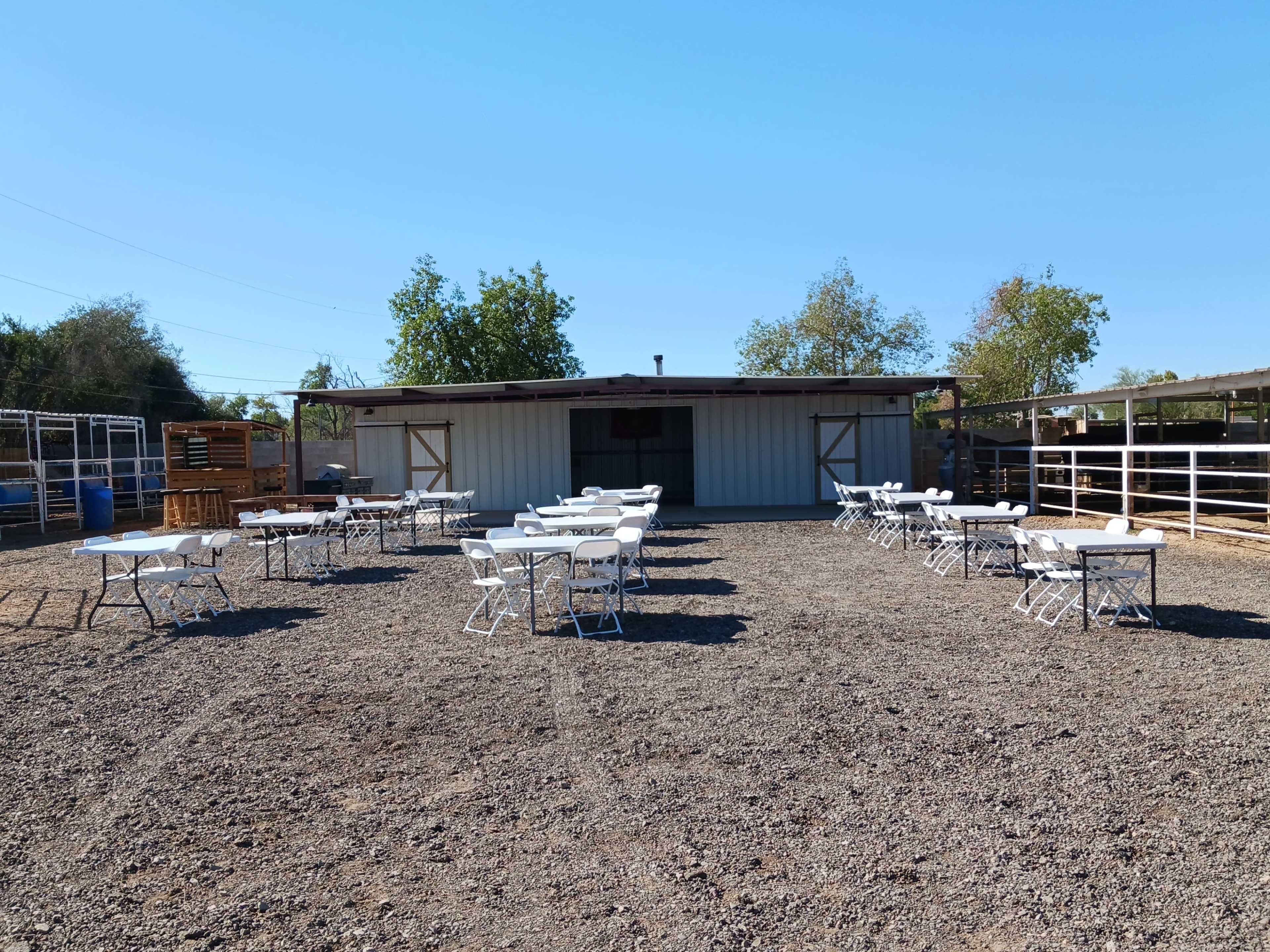 A gravel area with several white tables and chairs set up in front of a metal building, surrounded by trees and livestock enclosures.