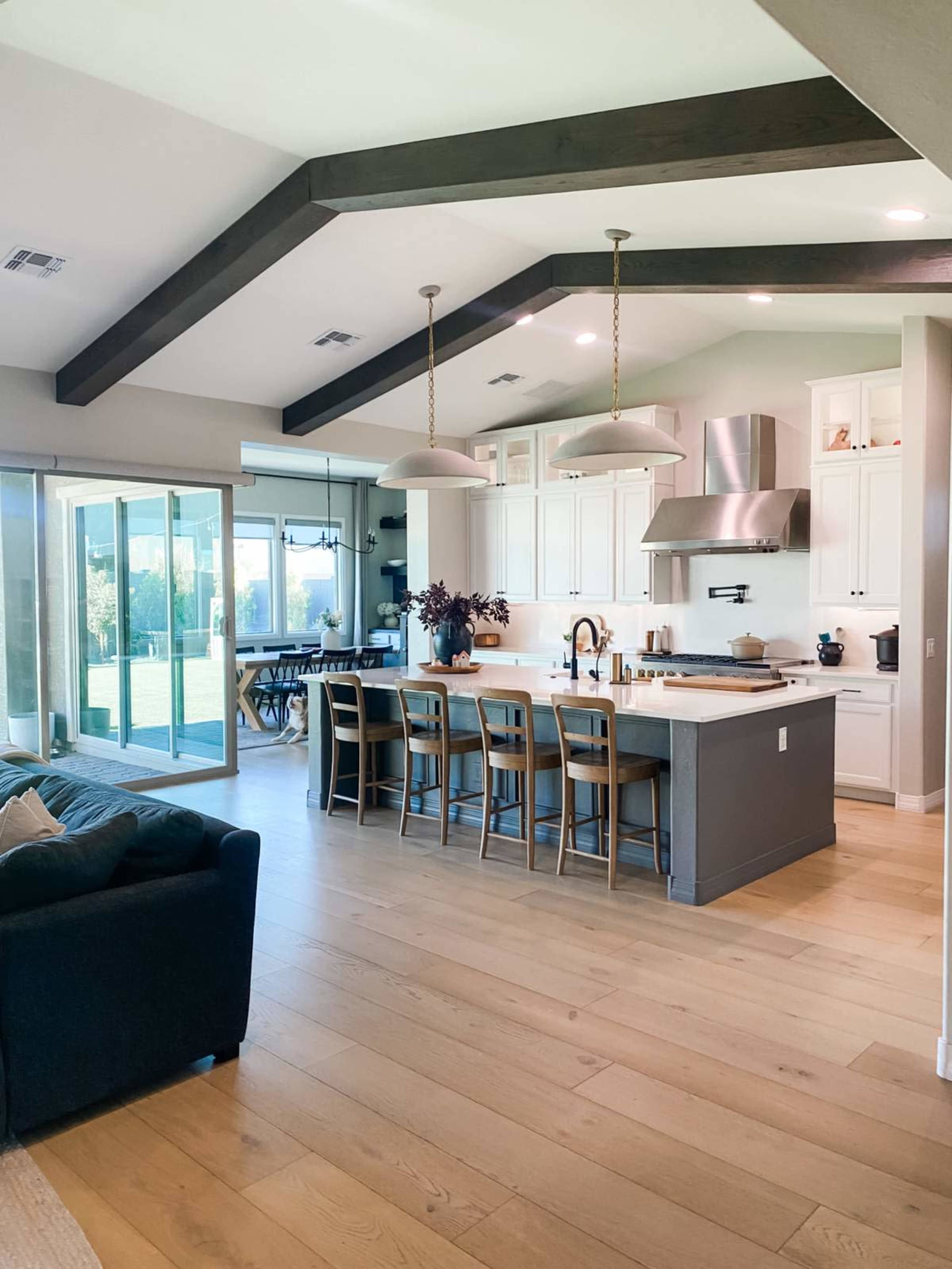 The image shows a modern kitchen with white cabinets, a large island, and a spacious open layout that connects to a living area with a dark sofa and large glass doors leading outside.