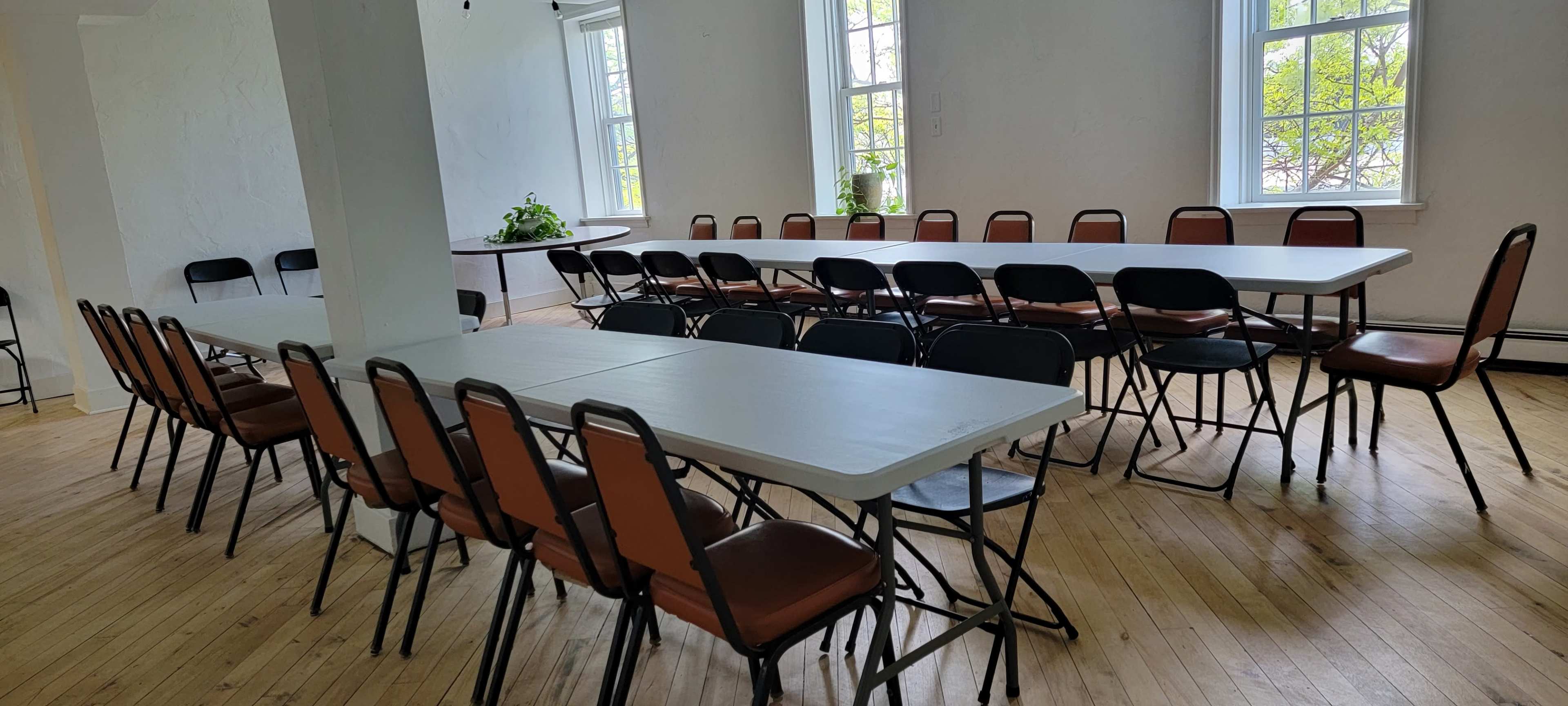 A spacious room features several long tables arranged in a U-shape, surrounded by chairs, with sunlight streaming through large windows.