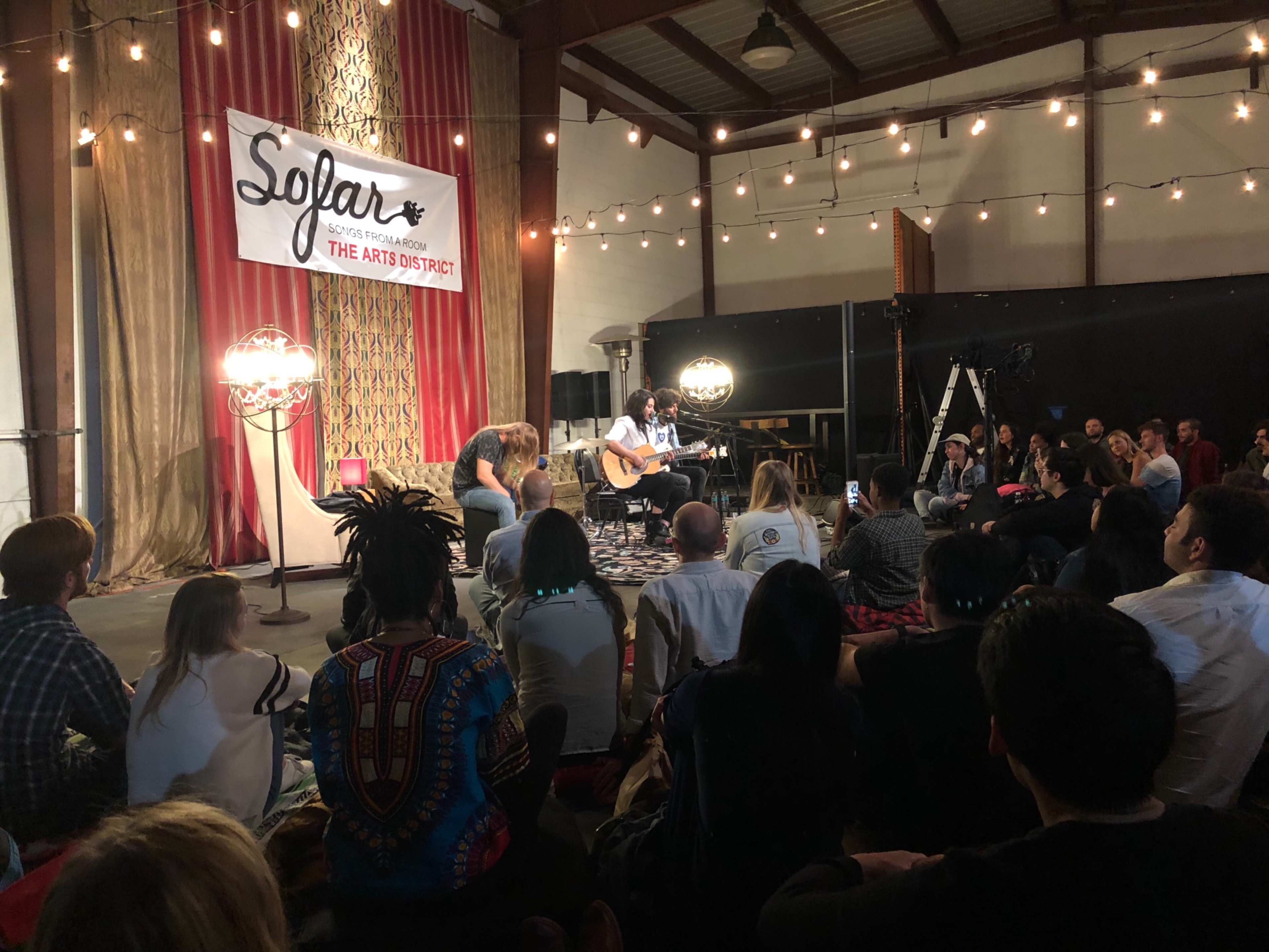 A musician performs with an acoustic guitar in a cozy venue filled with seated audience members under warm string lights.