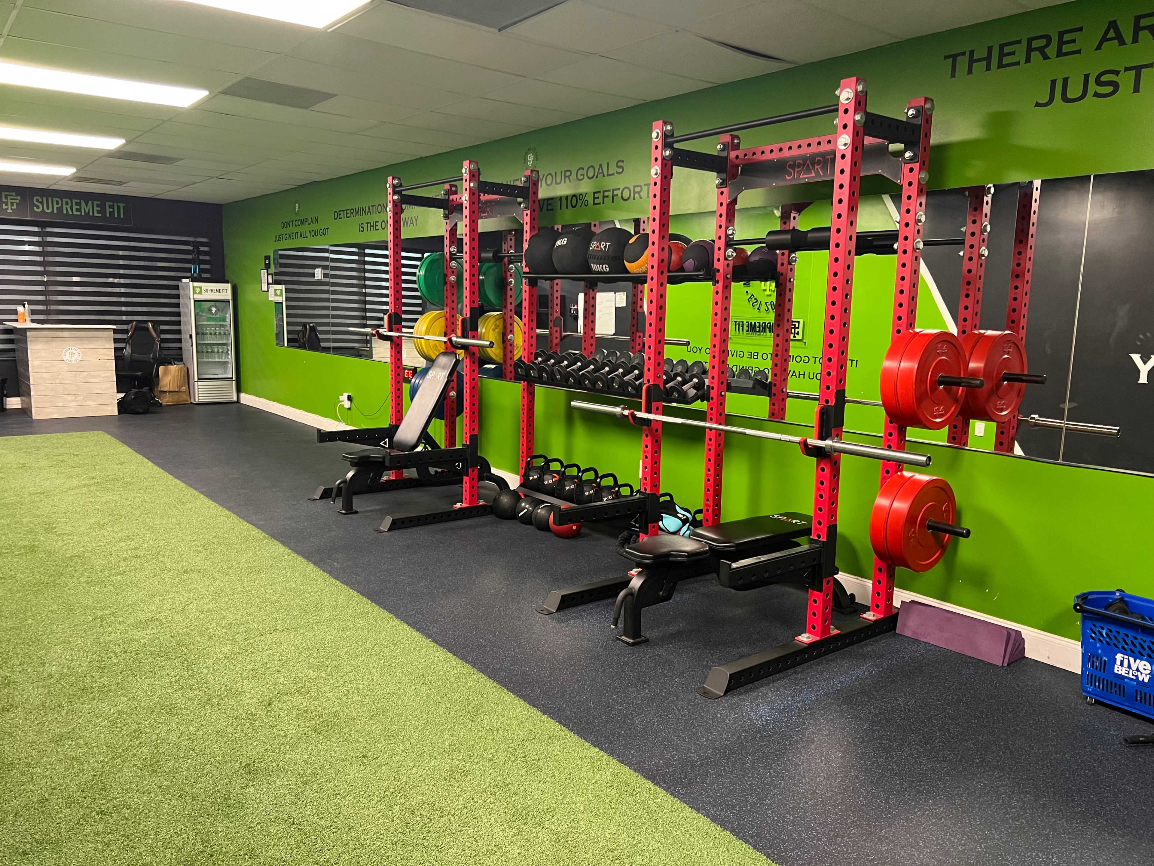 A gym area with weightlifting equipment, including benches, barbells, and free weights, against a green wall with motivational text.