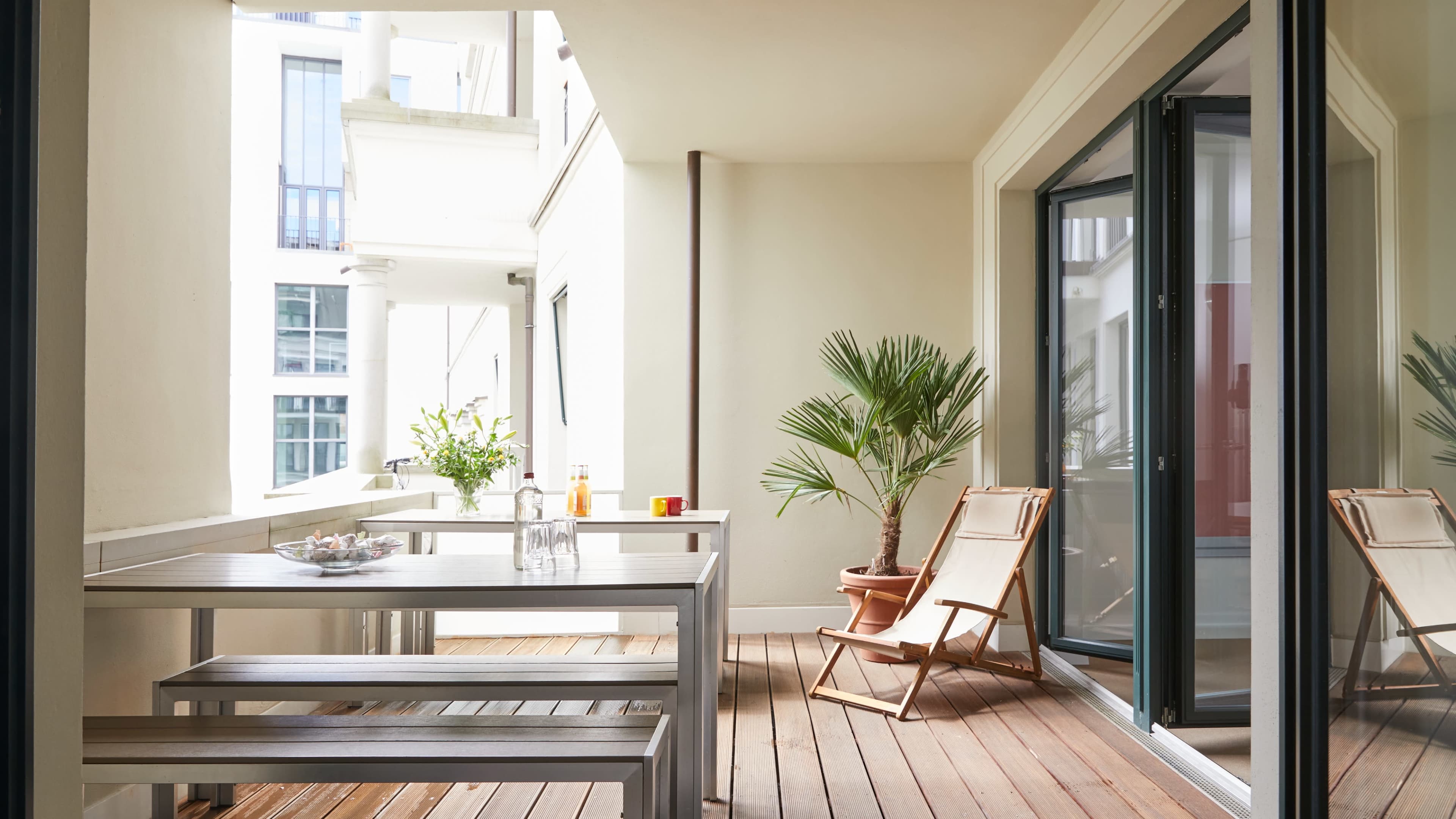 A modern balcony furnished with a wooden table, chairs, a potted plant, and a glass door leading to an interior space.