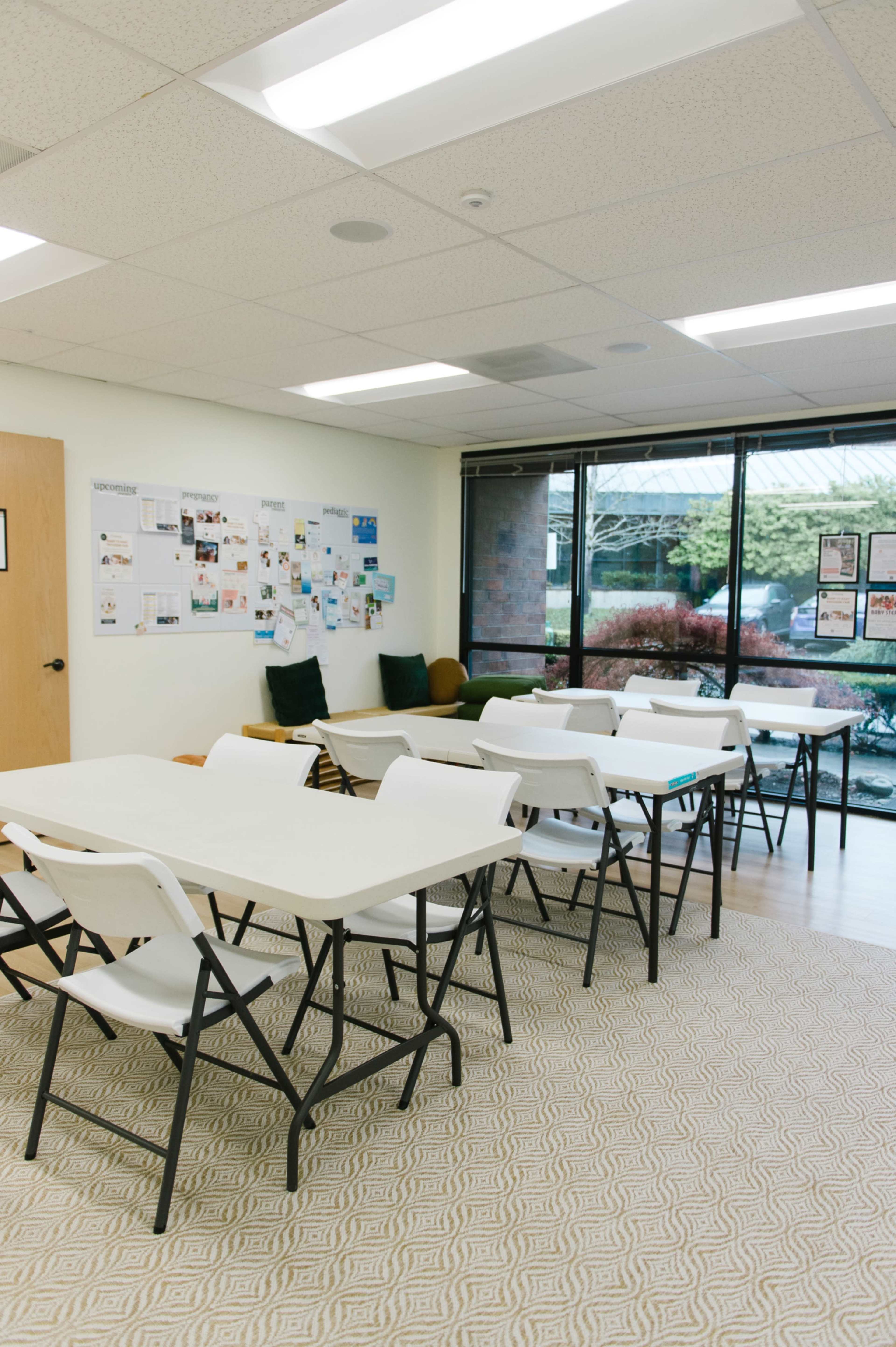 The image shows a well-lit classroom with several folding tables and chairs arranged facing a wall displaying various documents and images.
