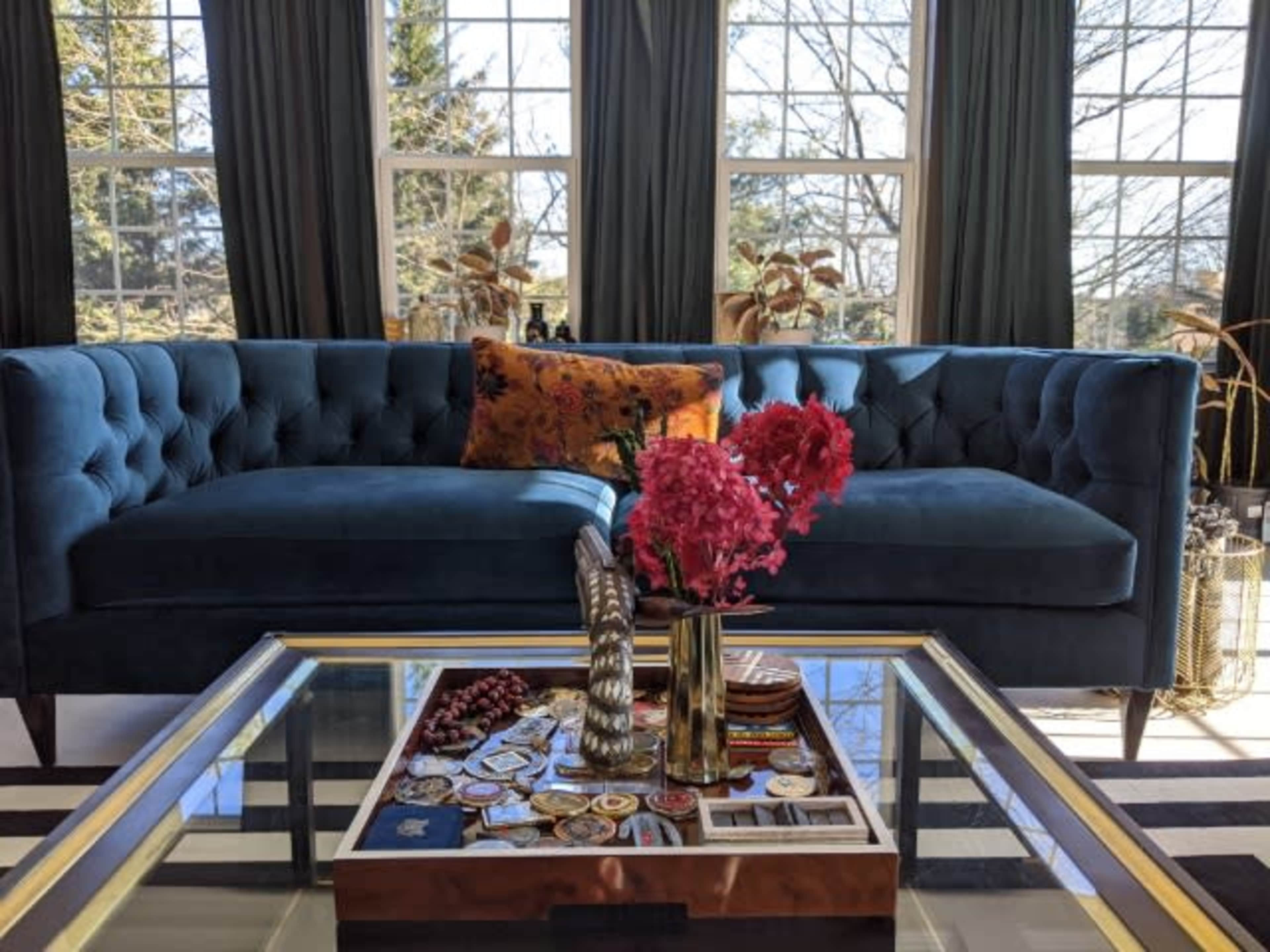 A blue tufted sofa with a decorative pillow, positioned in front of a coffee table adorned with flowers and various items, all set against large windows.
