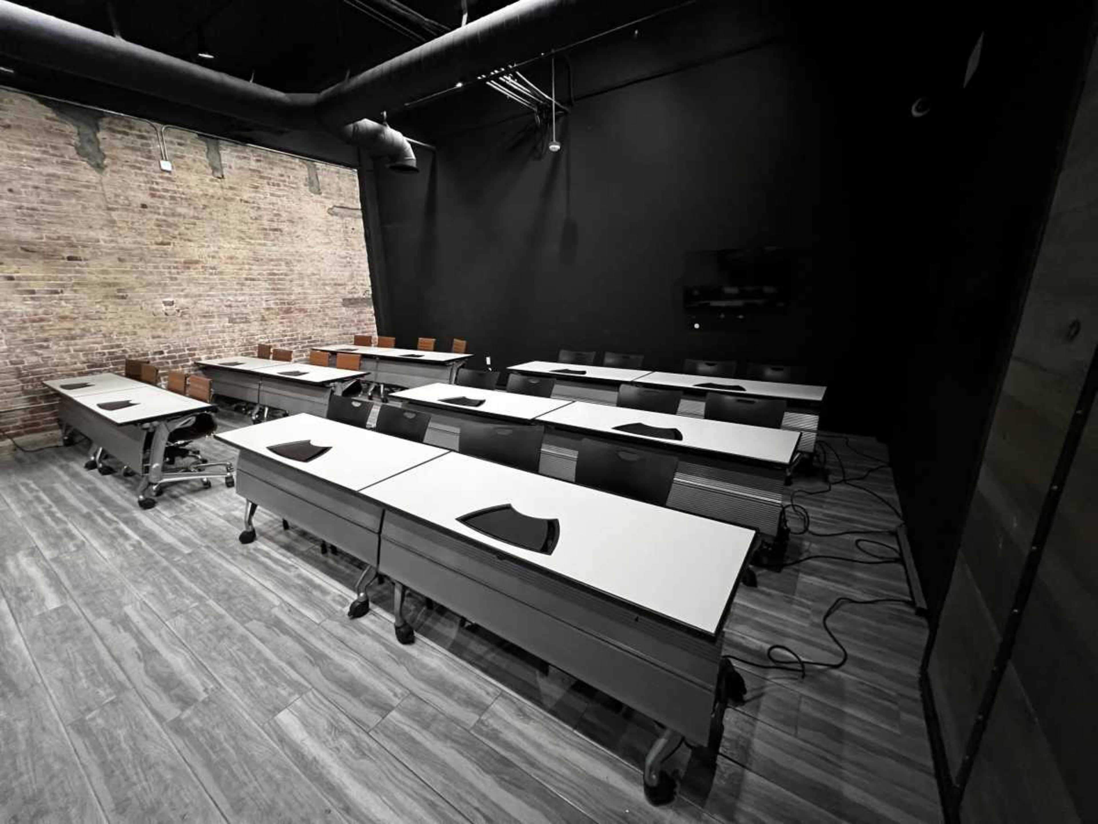 A modern classroom features multiple desks arranged in rows, with a black wall in the background and exposed brick on one side.