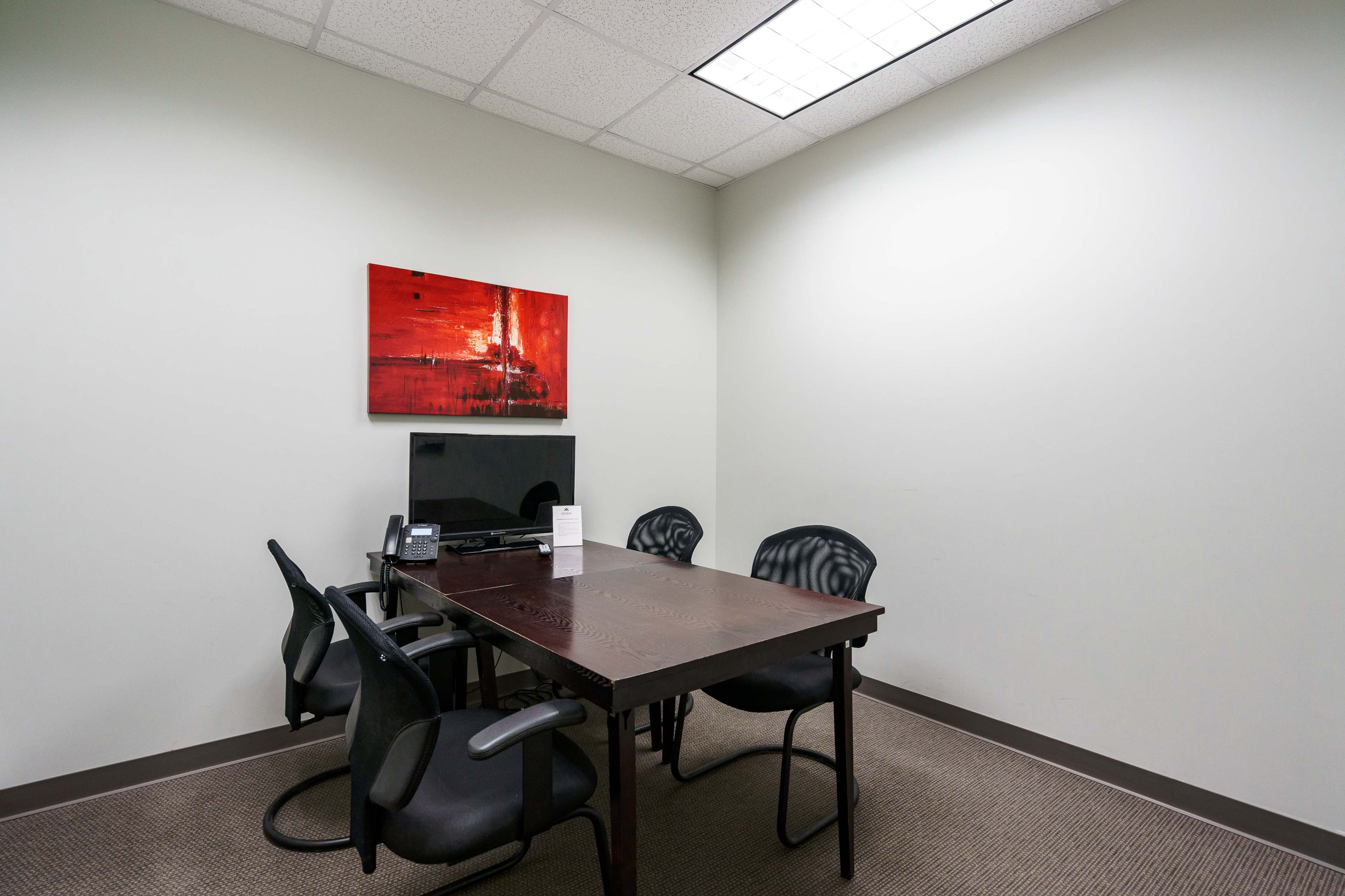 A small office meeting room containing a wooden table, four black chairs, a television on the wall, and a piece of abstract art.