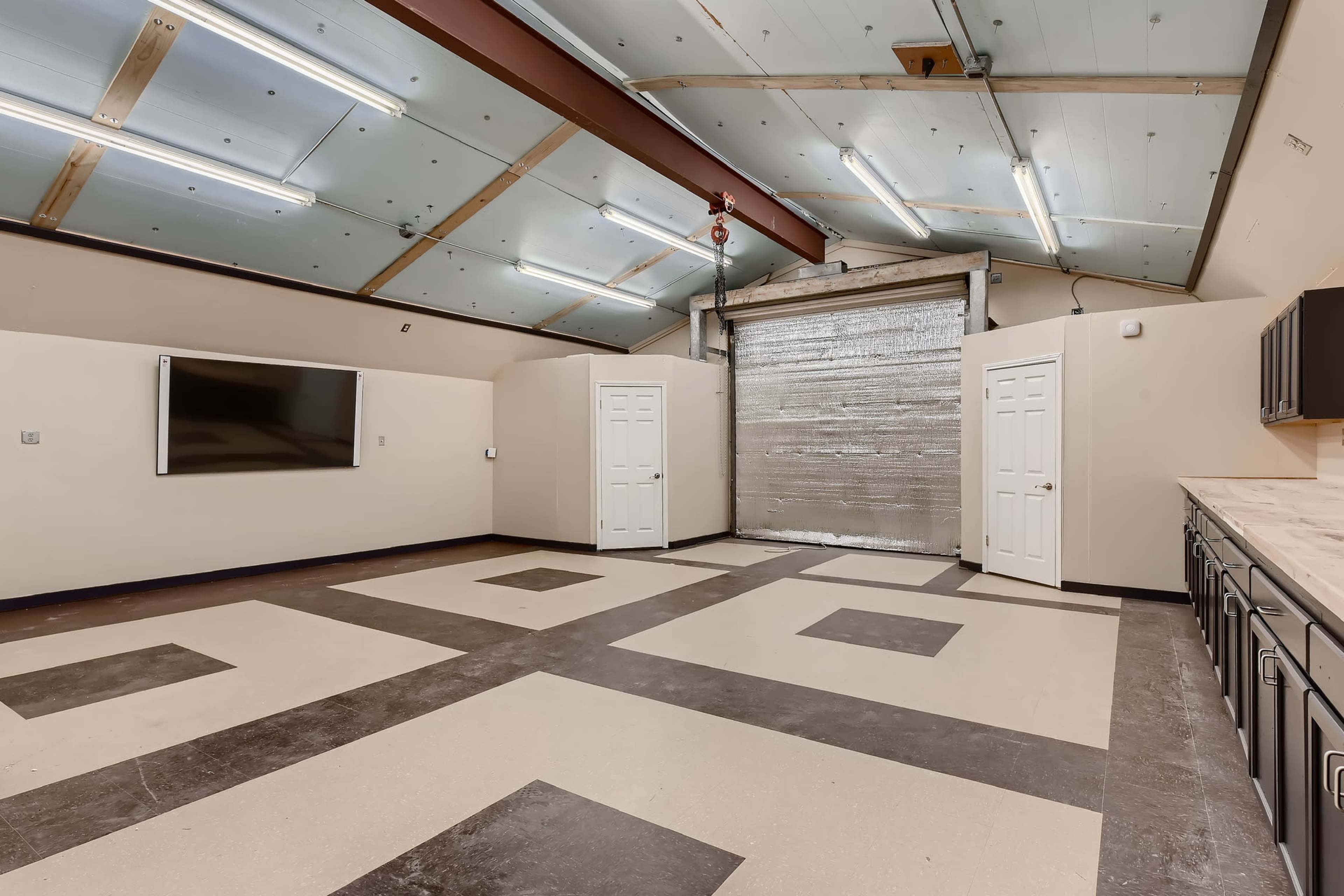 A spacious room with a large roll-up garage door, two white doors, a wall-mounted television, and a tiled floor featuring a geometric pattern.