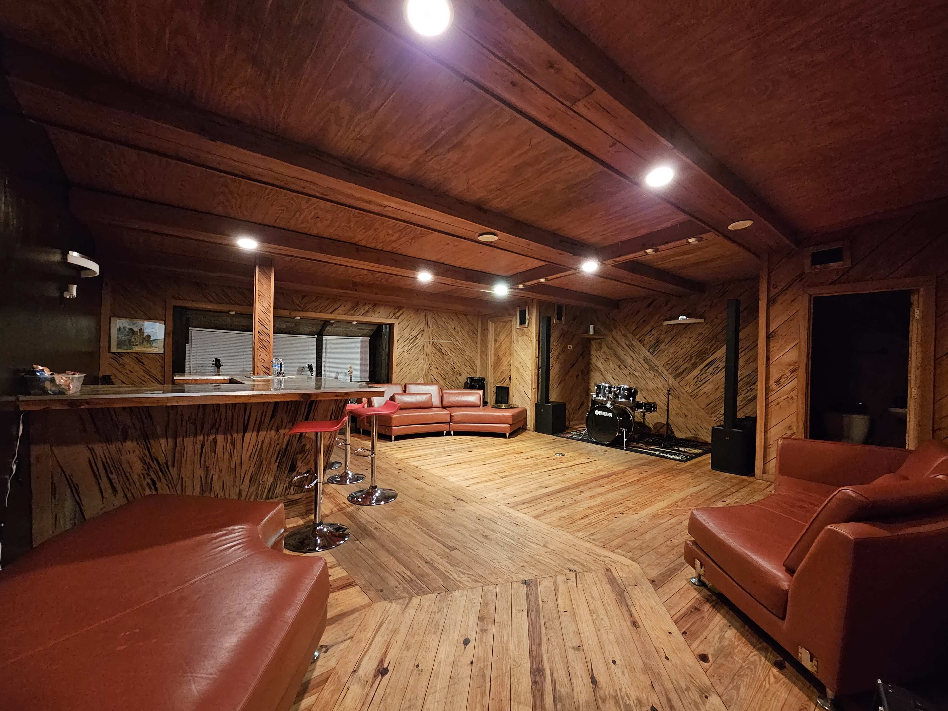 A spacious wooden-paneled room with a bar area, red leather seating, and a small stage equipped with musical instruments.