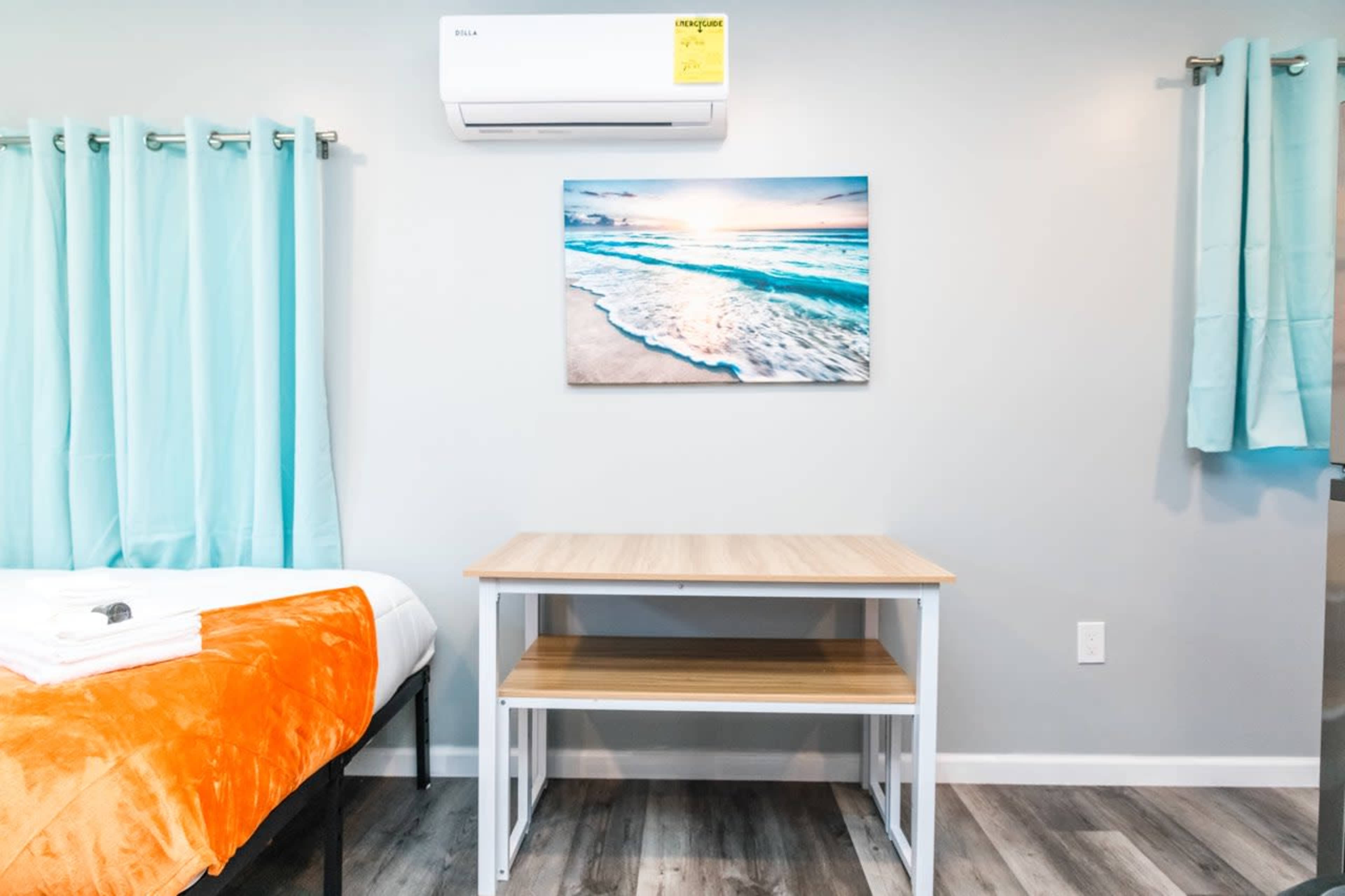 The image shows a room with a bed covered in an orange blanket, a wooden table against the wall, and a large ocean-themed picture above the table, with teal curtains on the window.