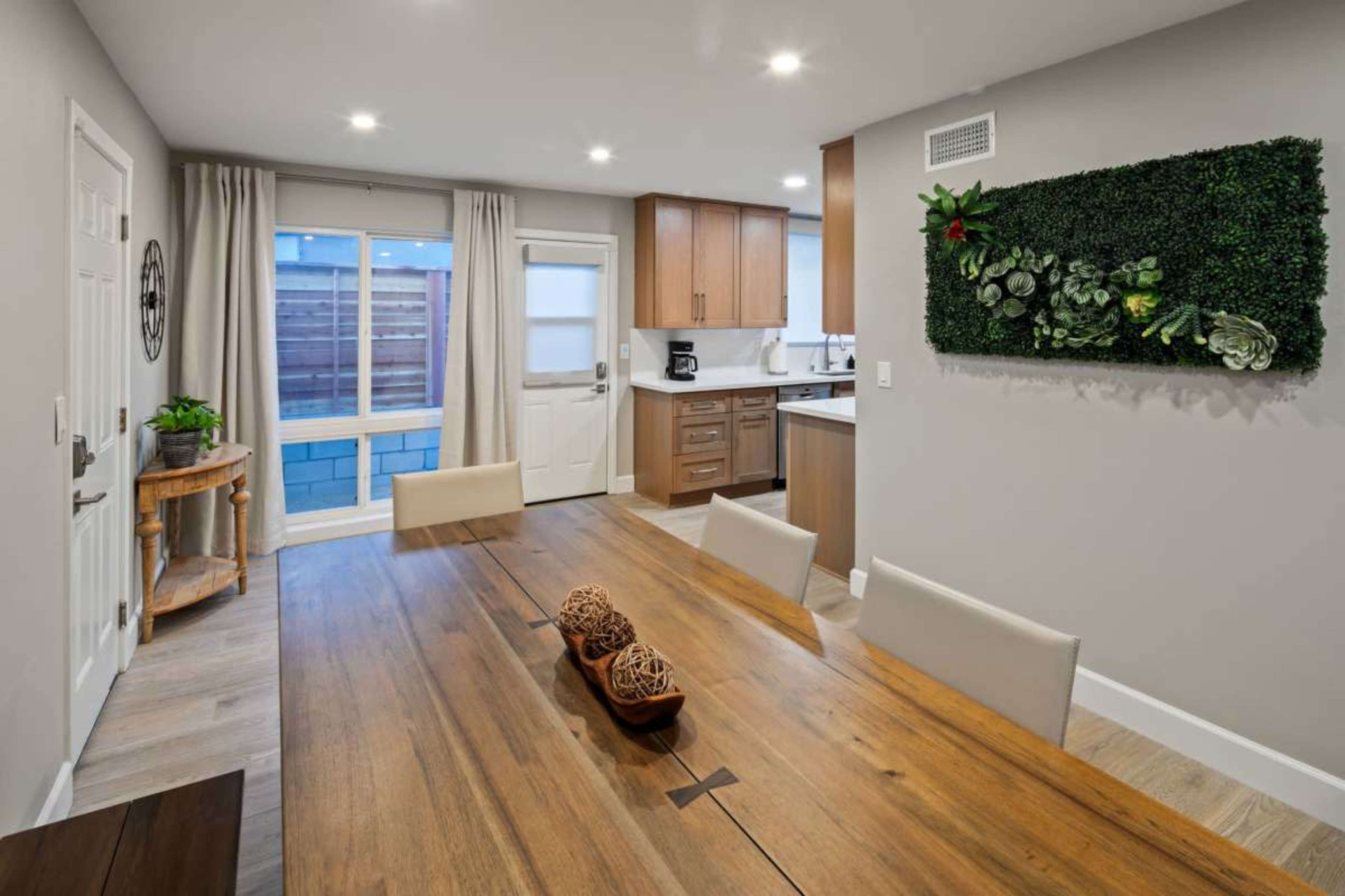 A modern dining area features a wooden table set with decorative elements, adjacent to a kitchen with wooden cabinets and a wall adorned with greenery.