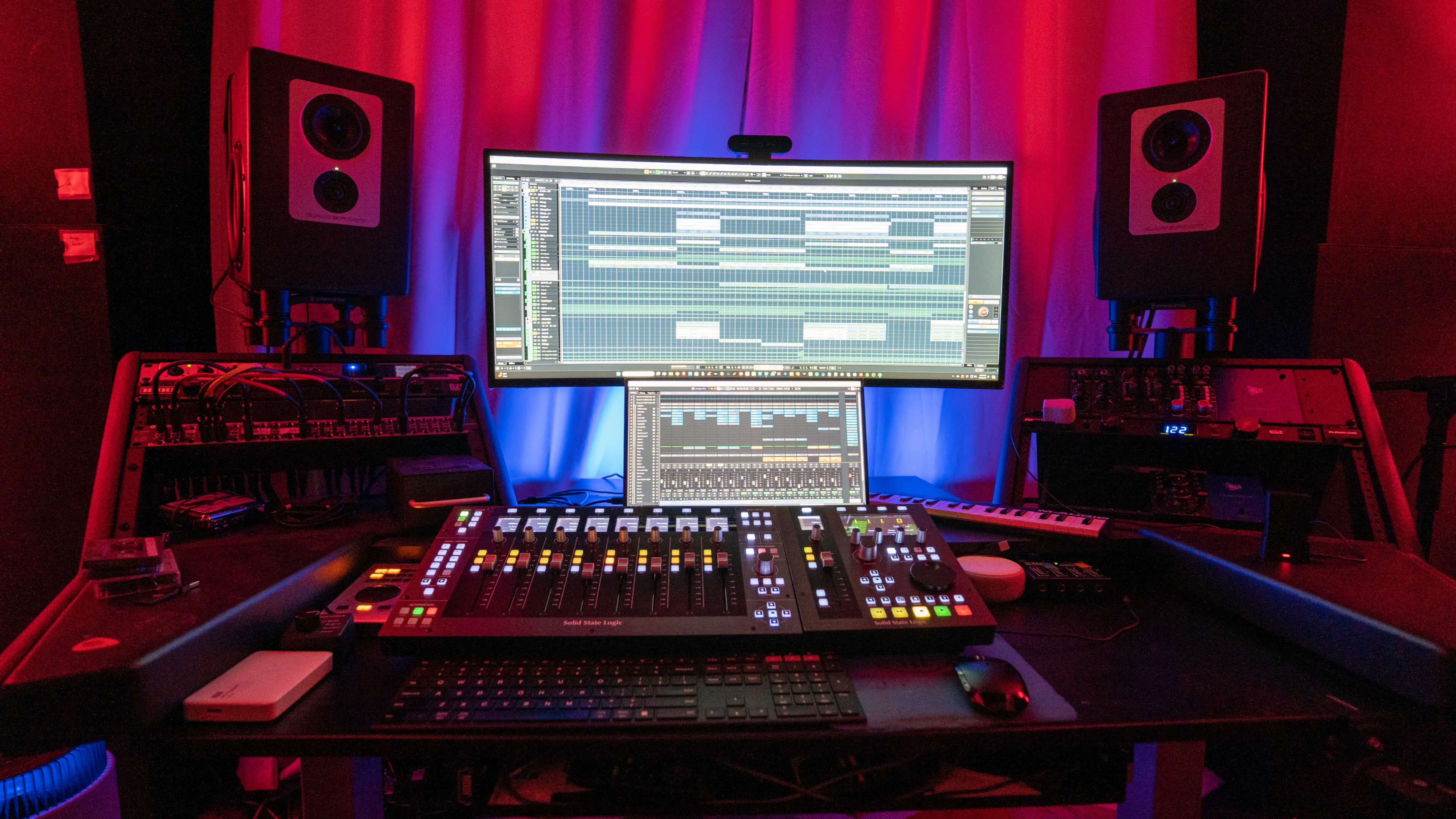 A music production studio featuring a large curved display, multiple audio monitors, and a mixing console with various controls and a computer setup.