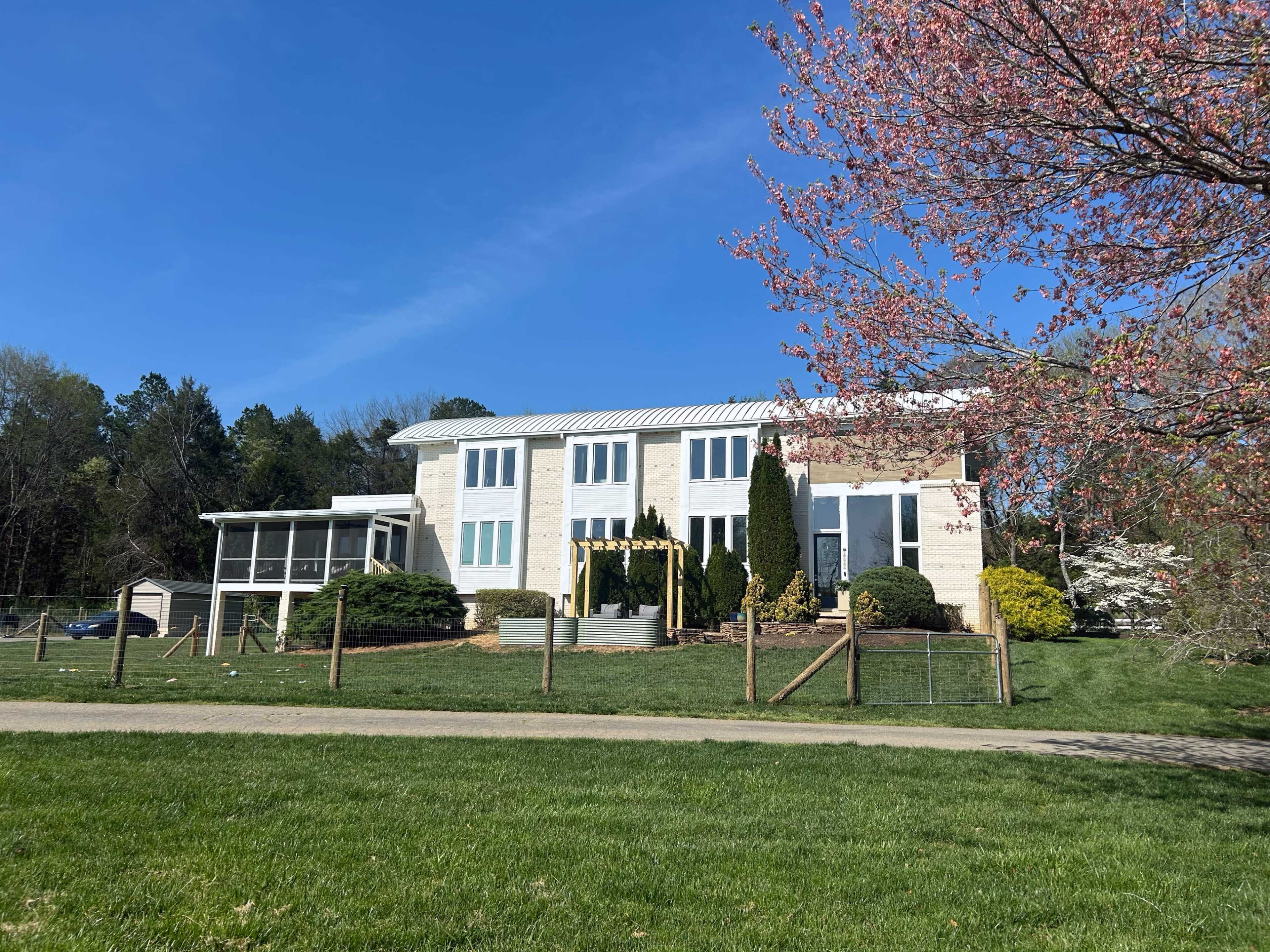 A modern two-story house with a landscaped yard and a covered porch is surrounded by trees and a clear blue sky.