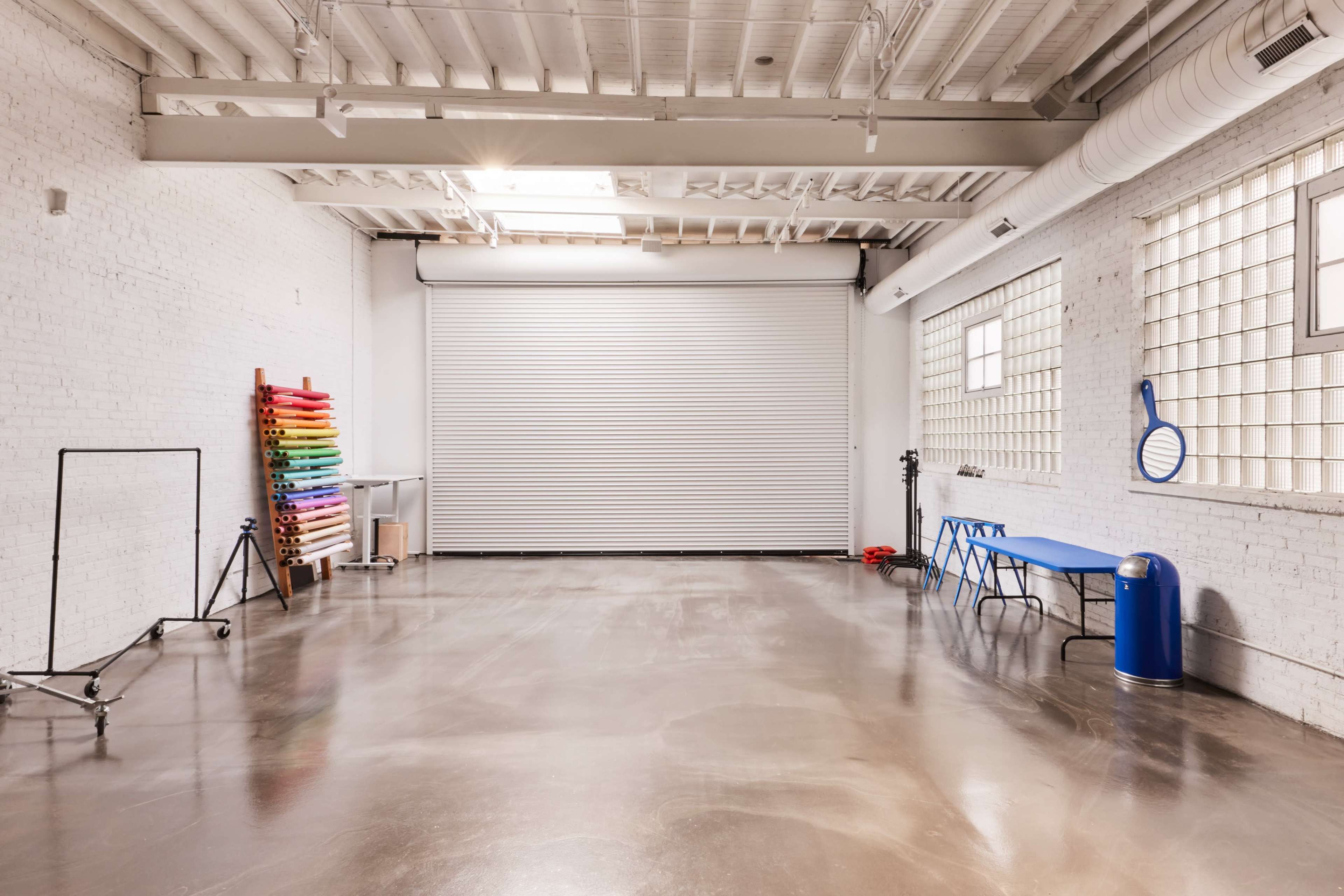 A spacious, minimalistic interior with a roll-up door, a blue table, and a rainbow-colored backdrop stand against a white brick wall.