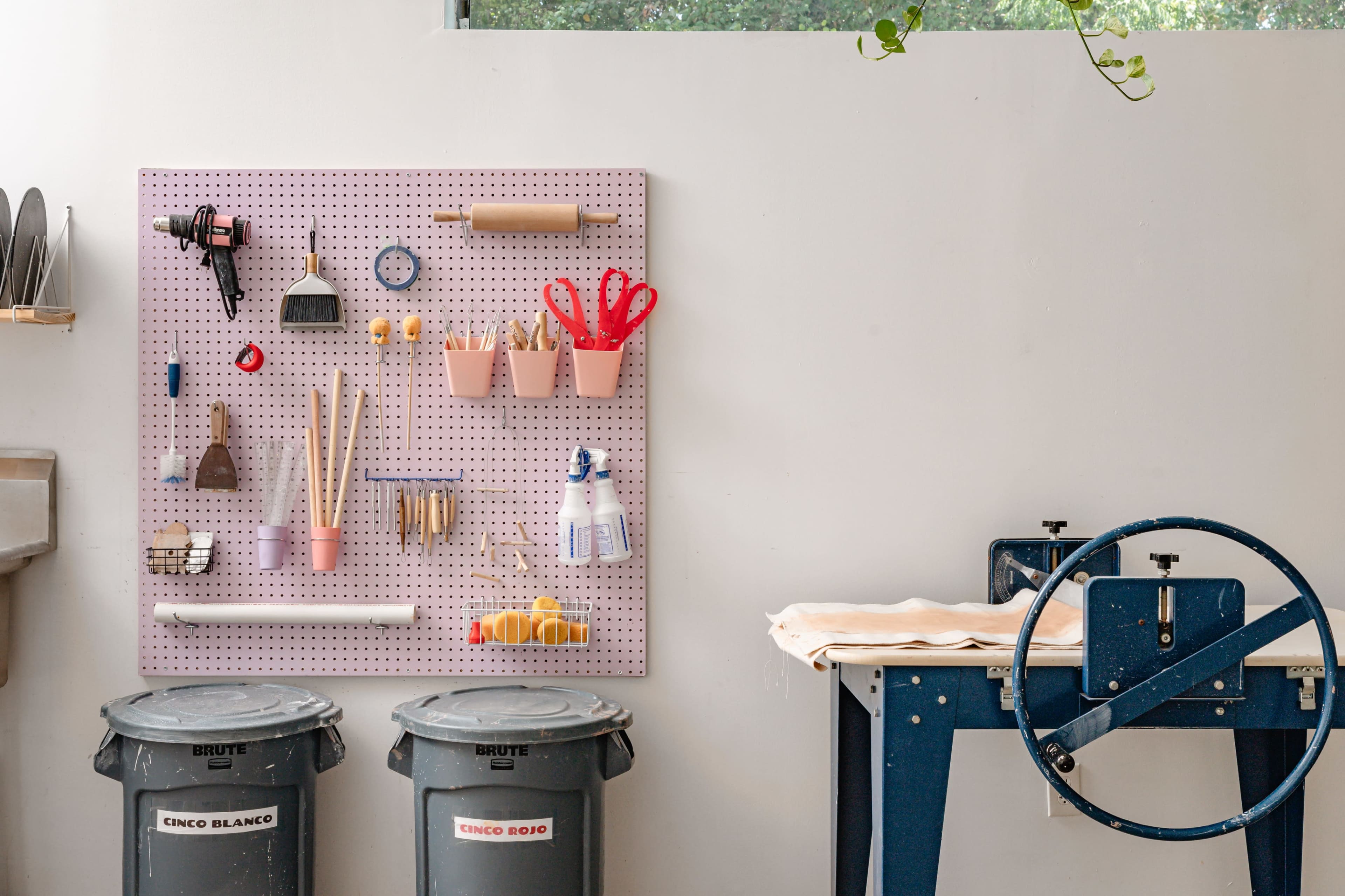 A pegboard with various tools and supplies is mounted on a wall above two trash bins and a work table.