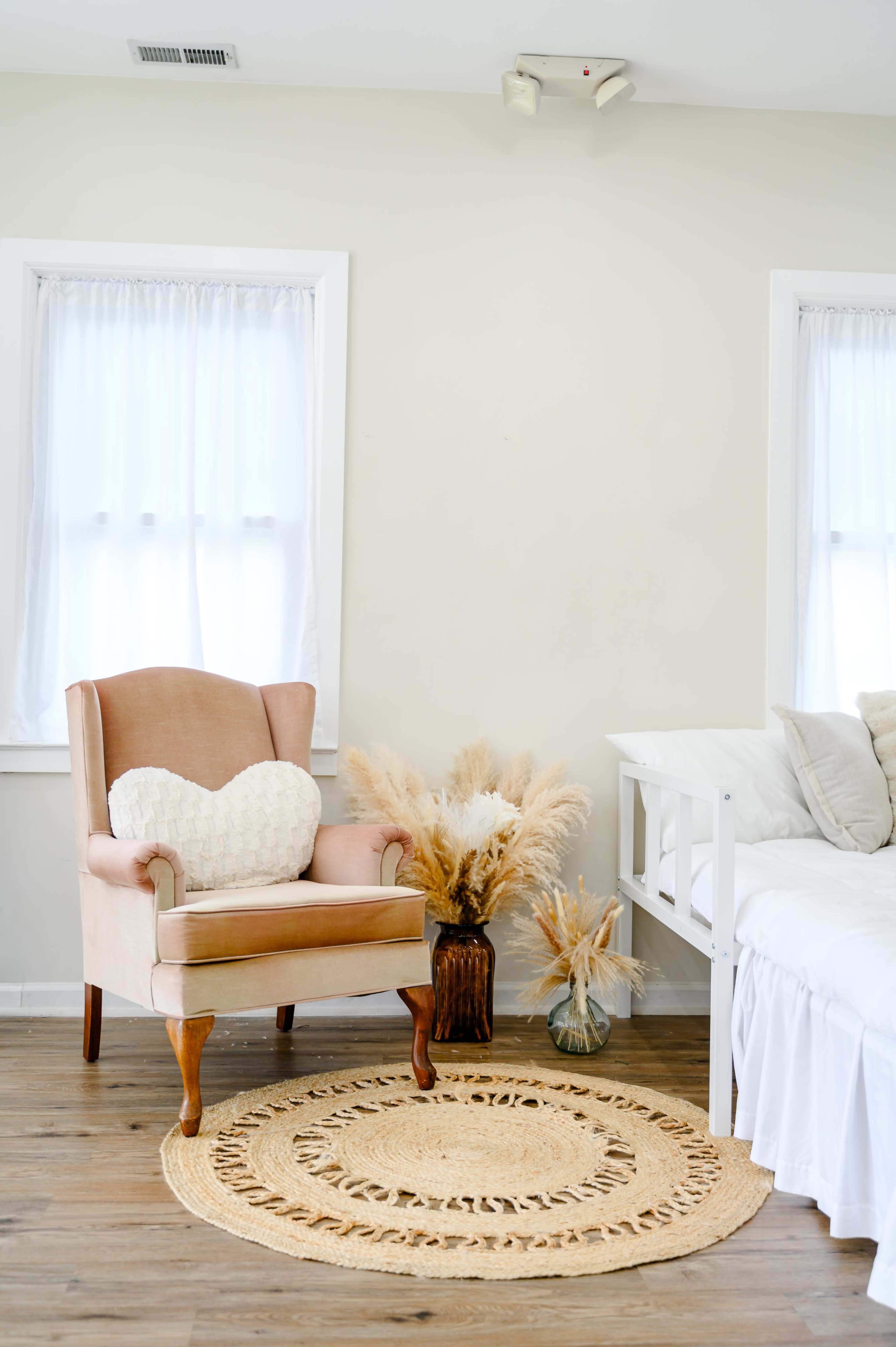 A pink upholstered armchair with a heart-shaped cushion is positioned next to a decorative arrangement of dried plants in a light-colored room.