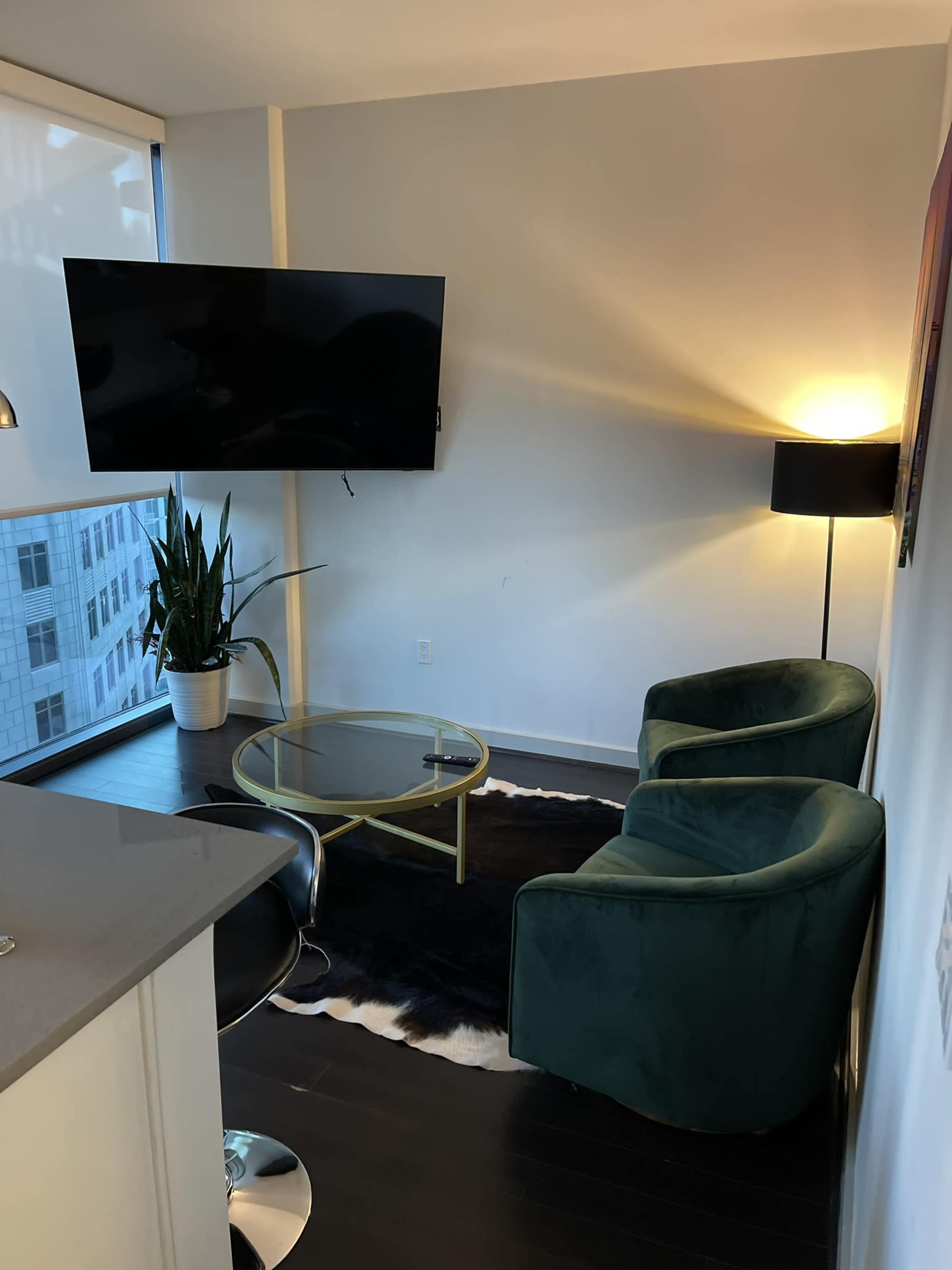 A modern living space features two green chairs, a glass coffee table on a black and white rug, a floor lamp, and a television mounted on the wall beside a large window.