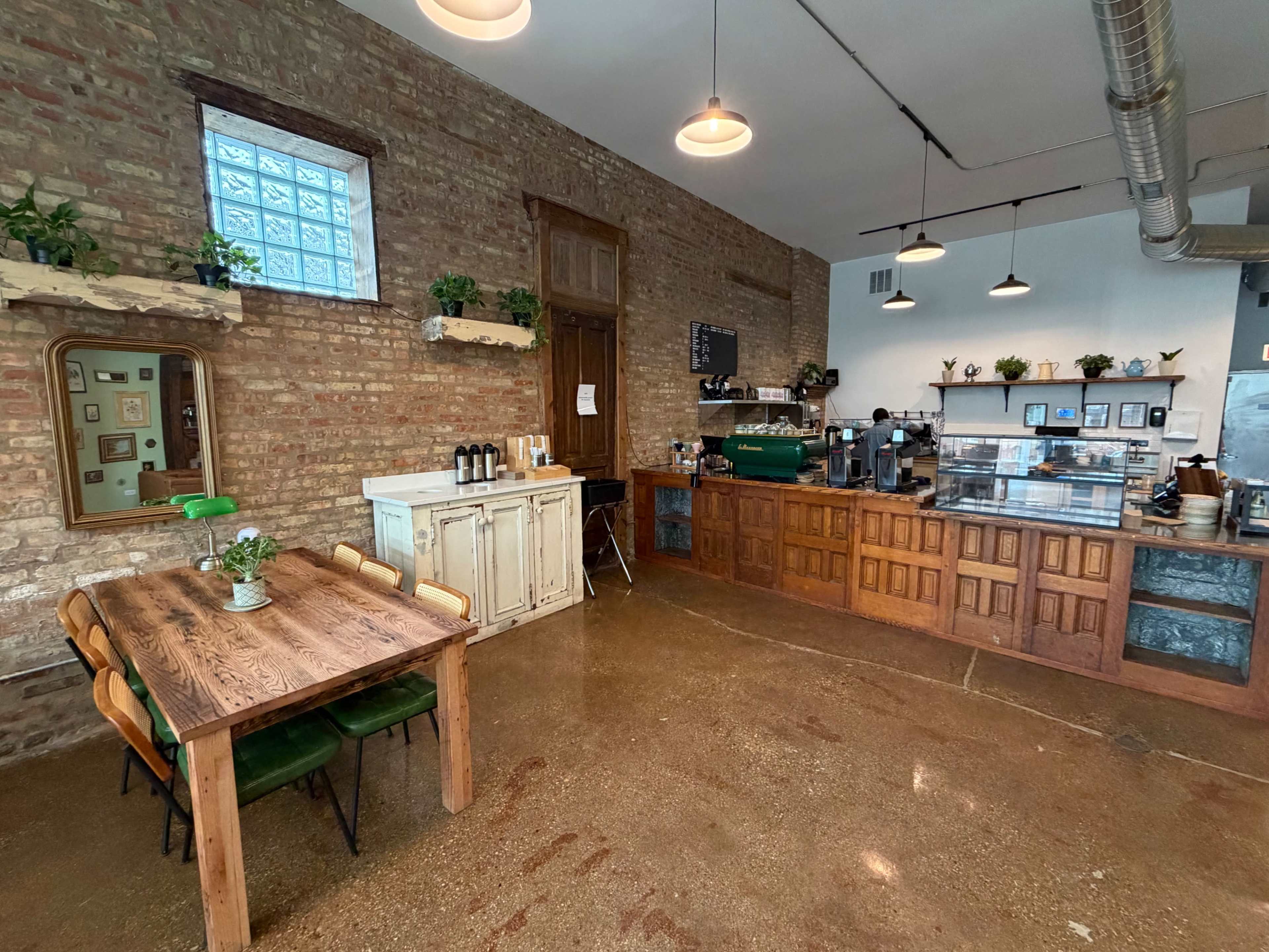 The image shows a modern café interior with exposed brick walls, a wooden service counter, and a rustic dining table with green chairs.