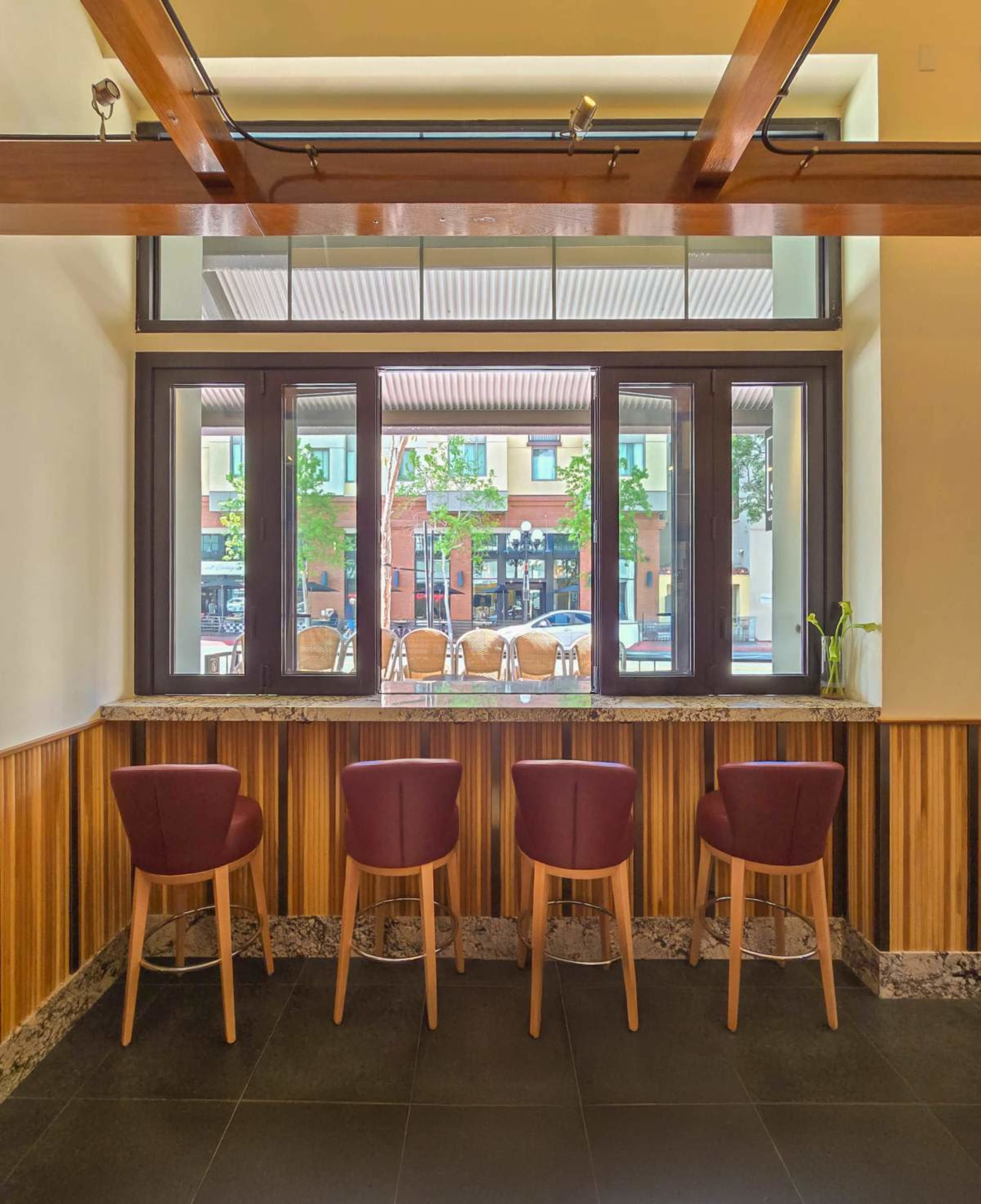 The image shows a modern interior with four red-chairs placed at a marble countertop against a backdrop of large windows overlooking an outdoor seating area.