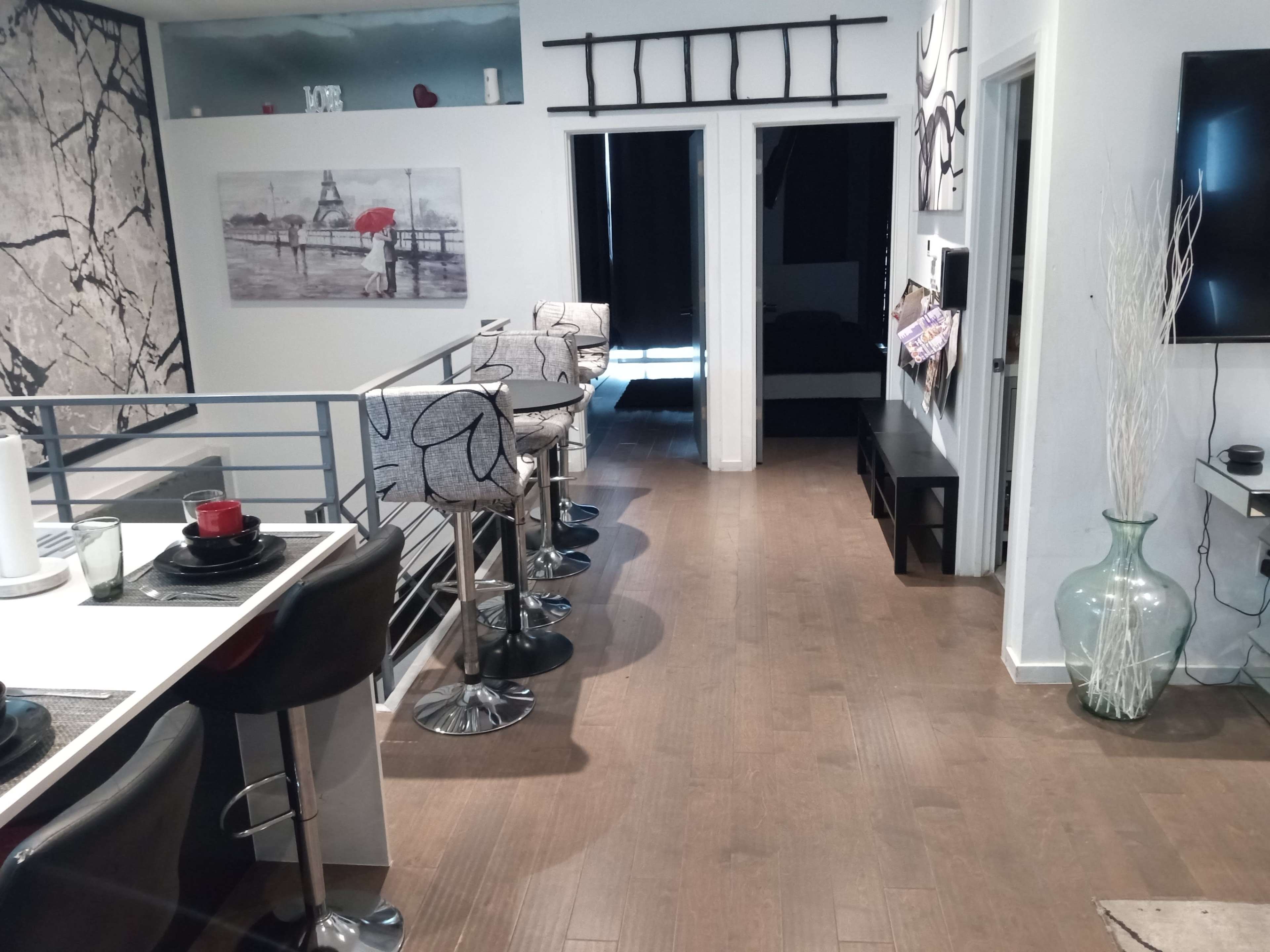 The image displays a modern interior with a dining area featuring high stools, a hallway leading to two doorways, and a decorative wall with artwork.