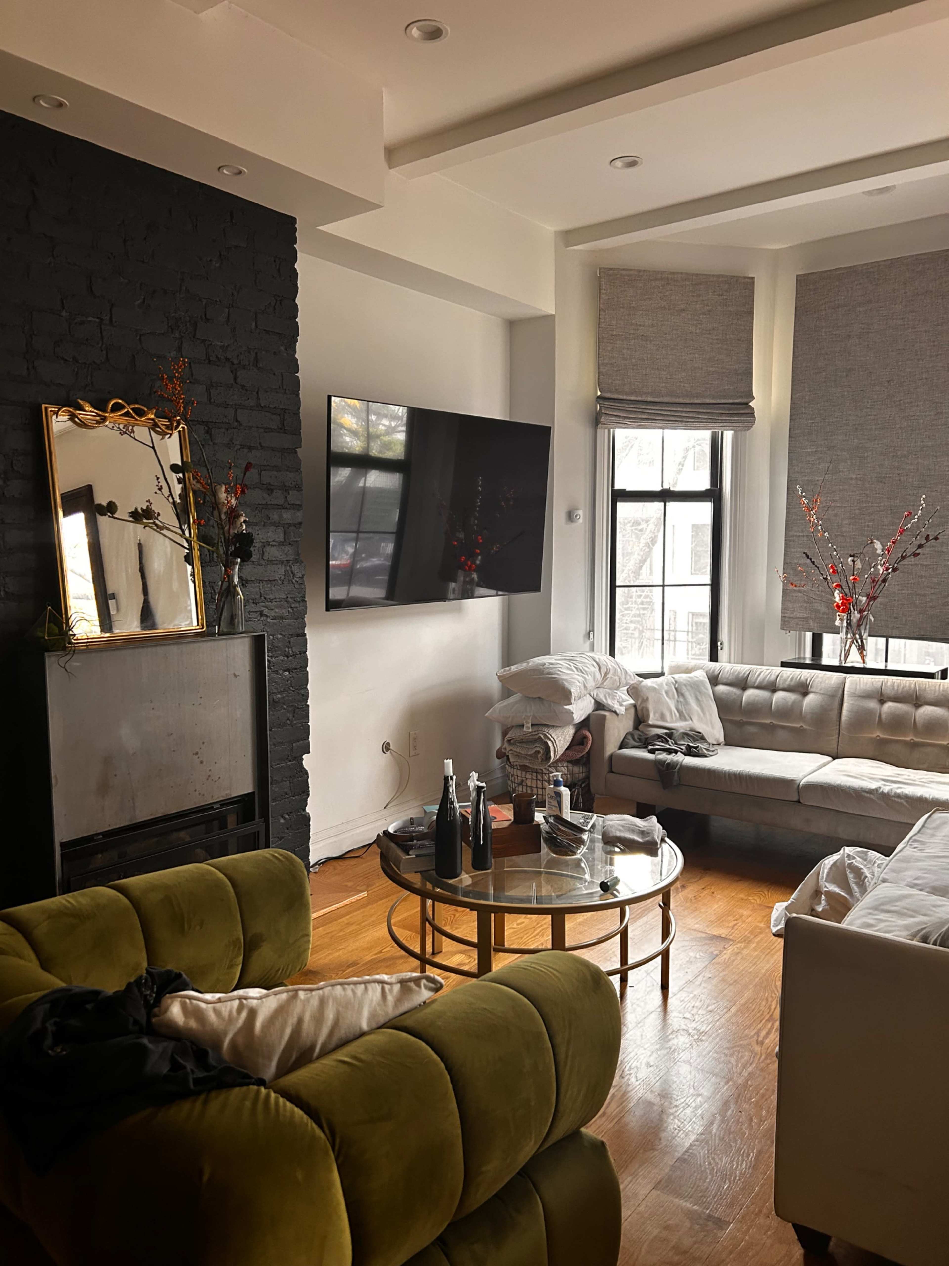 The image shows a modern living room featuring a green sofa, a round coffee table with drinks, a large wall-mounted TV, and large windows with shades.