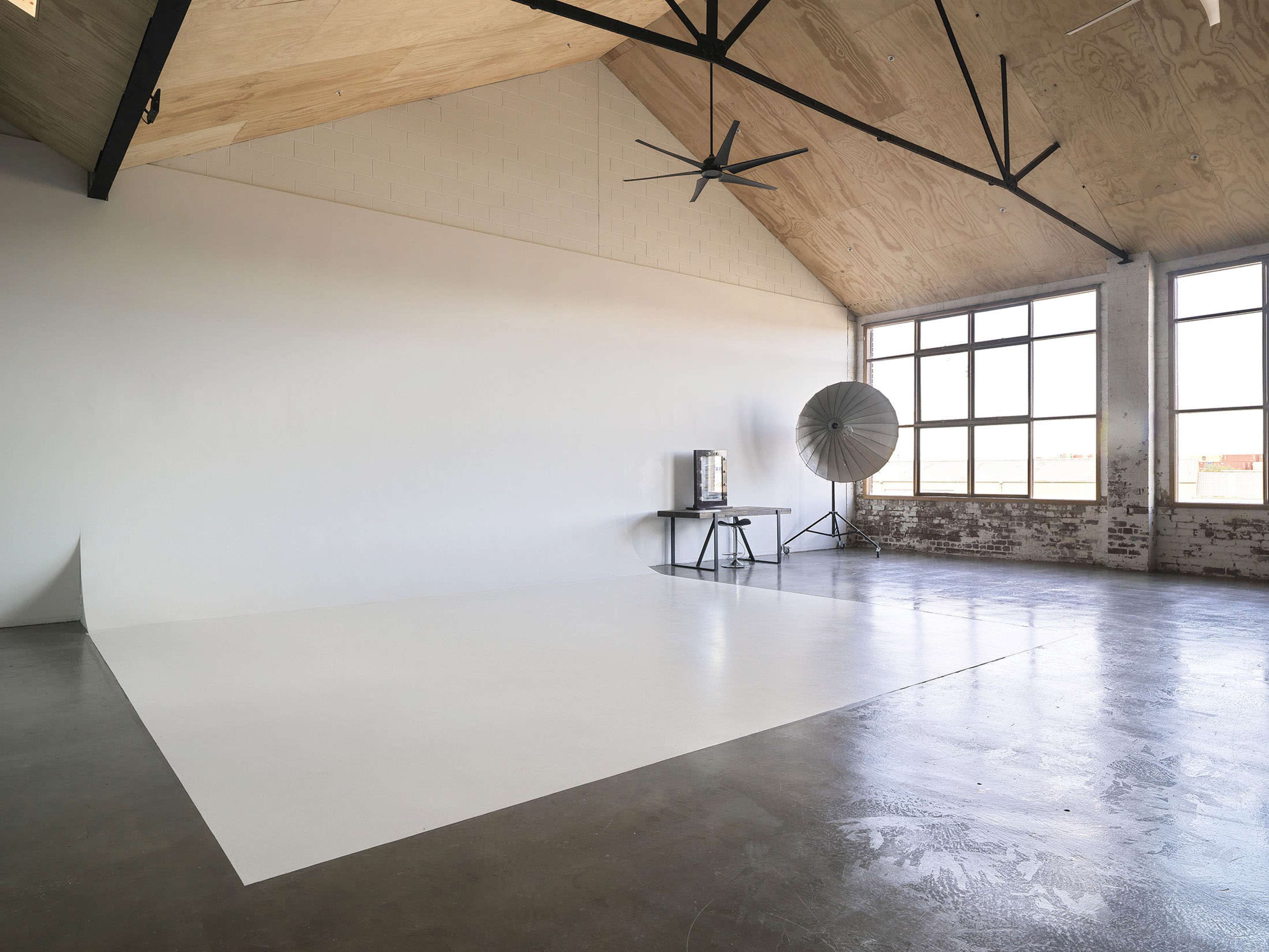 A spacious indoor studio featuring a large white wall, concrete flooring, a table with a coffee maker, and a large circular fan near a set of windows.