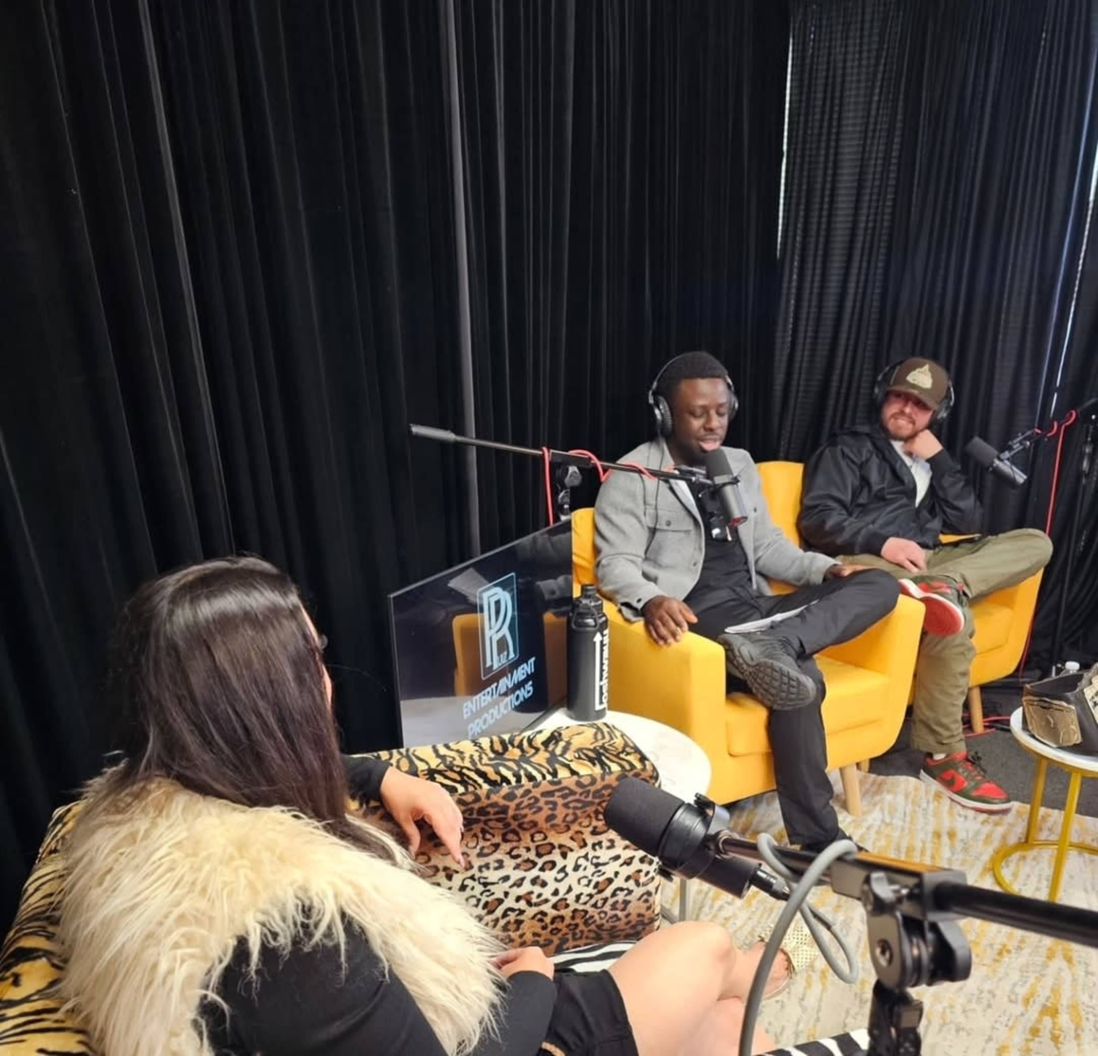Three people are sitting in a studio setting with microphones, engaged in a discussion, while two of them are on yellow chairs and one is in a leopard-print chair.