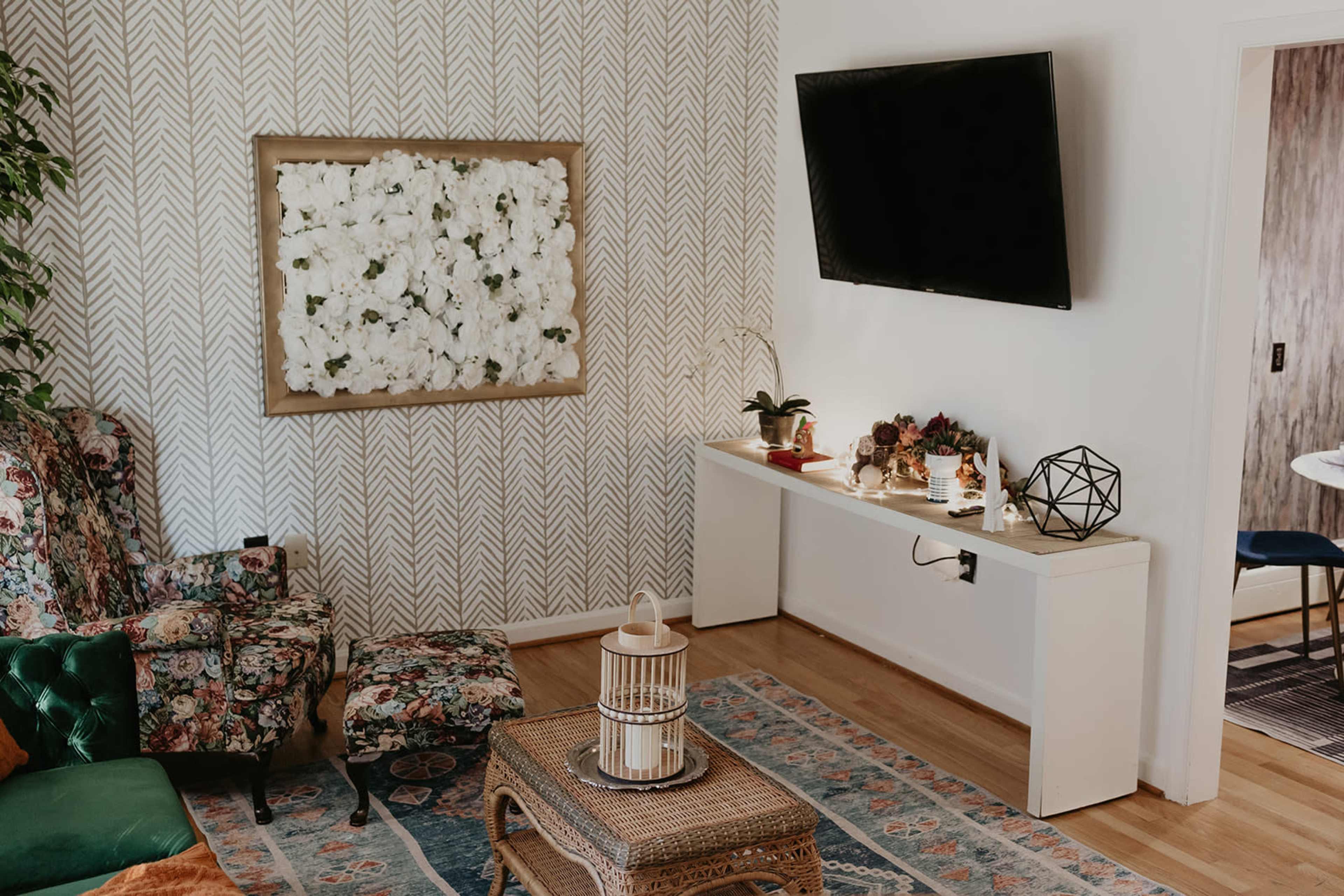 A cozy living room with floral-patterned furniture, a wall-mounted TV, and a console table decorated with plants and decorative objects.