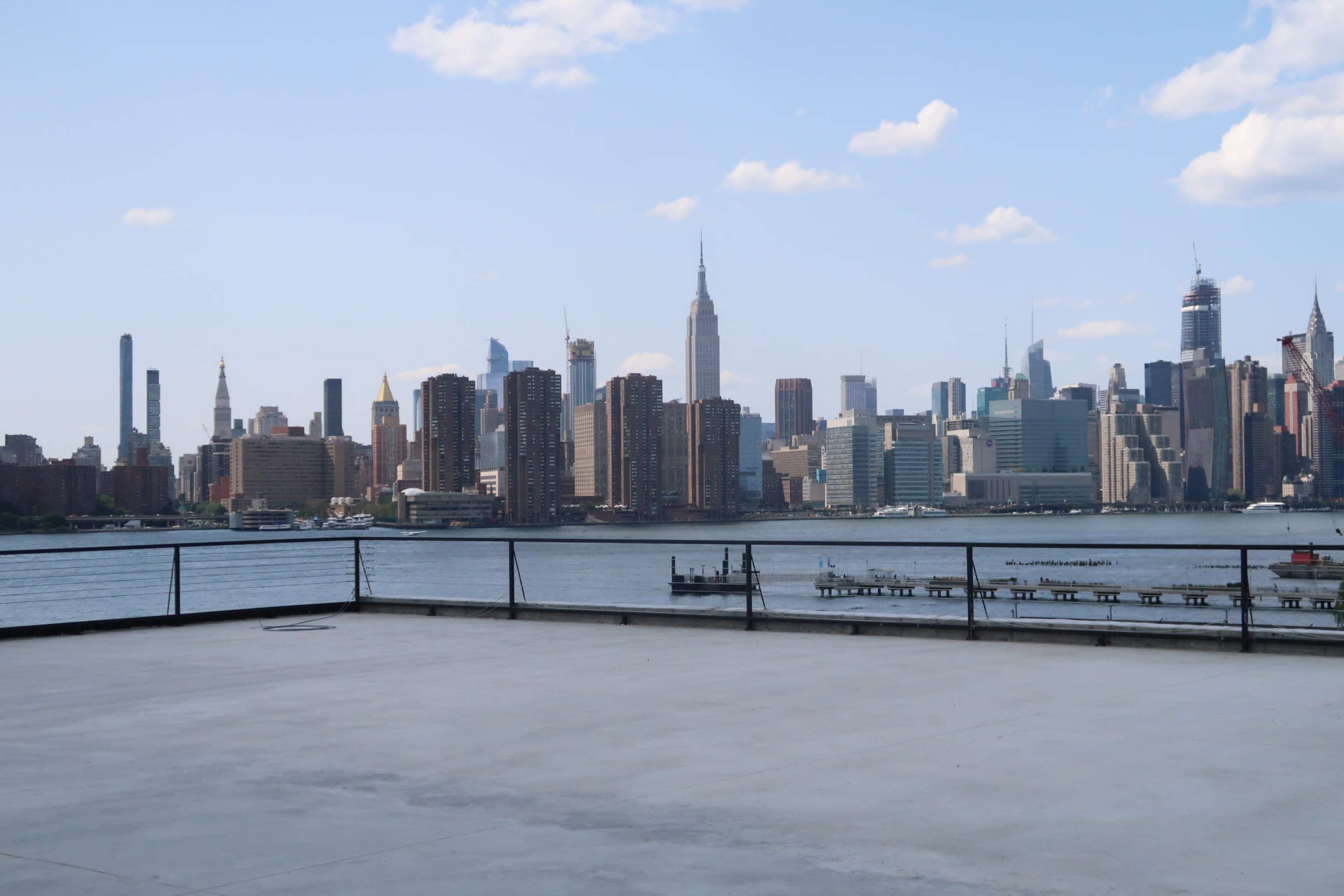 A wide view of the New York City skyline from a waterfront location, with the Empire State Building prominently visible among other skyscrapers.