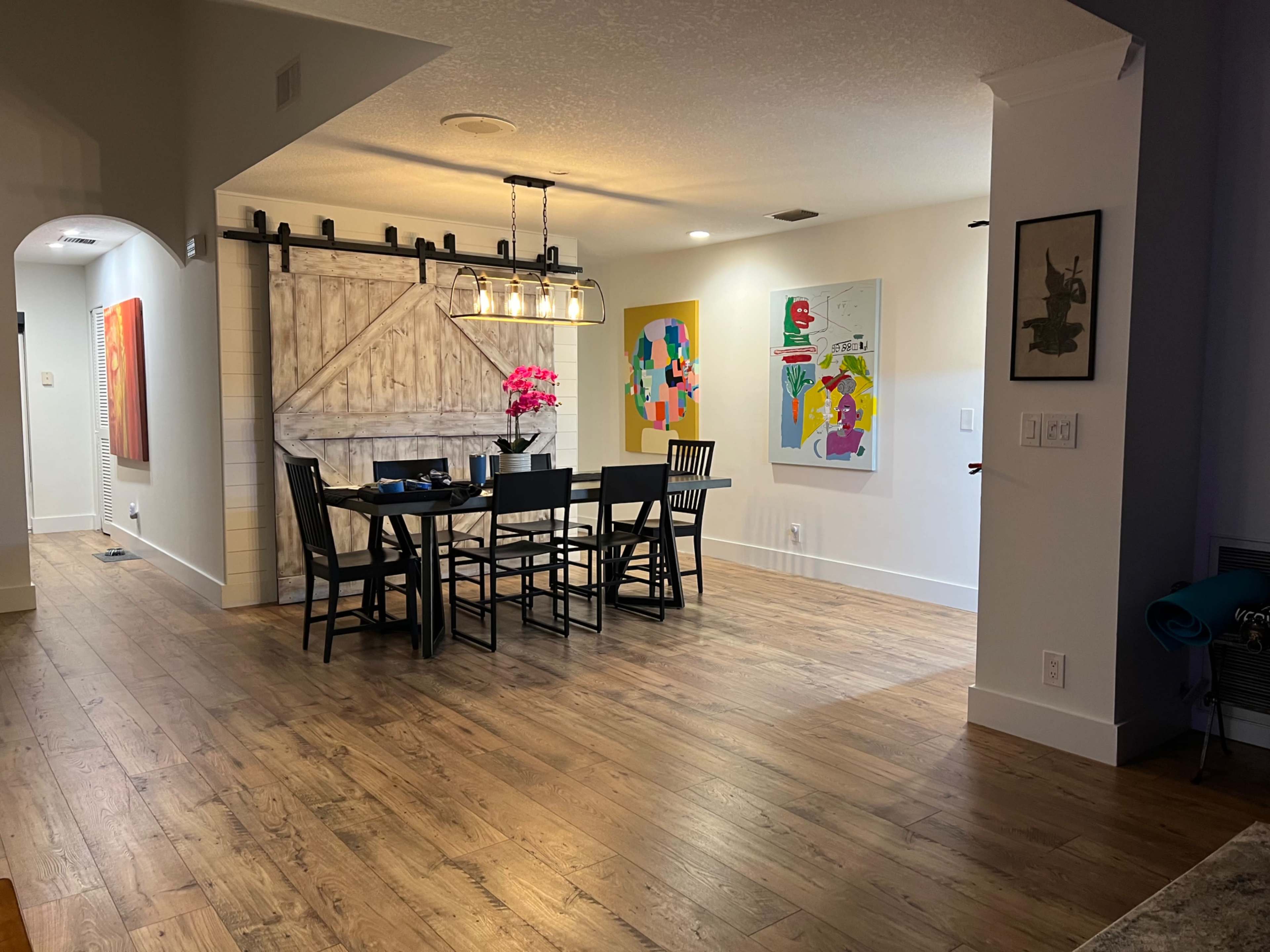 The image shows a dining area with a dark wooden table and black chairs, illuminated by a pendant light, and features colorful artwork on the walls.