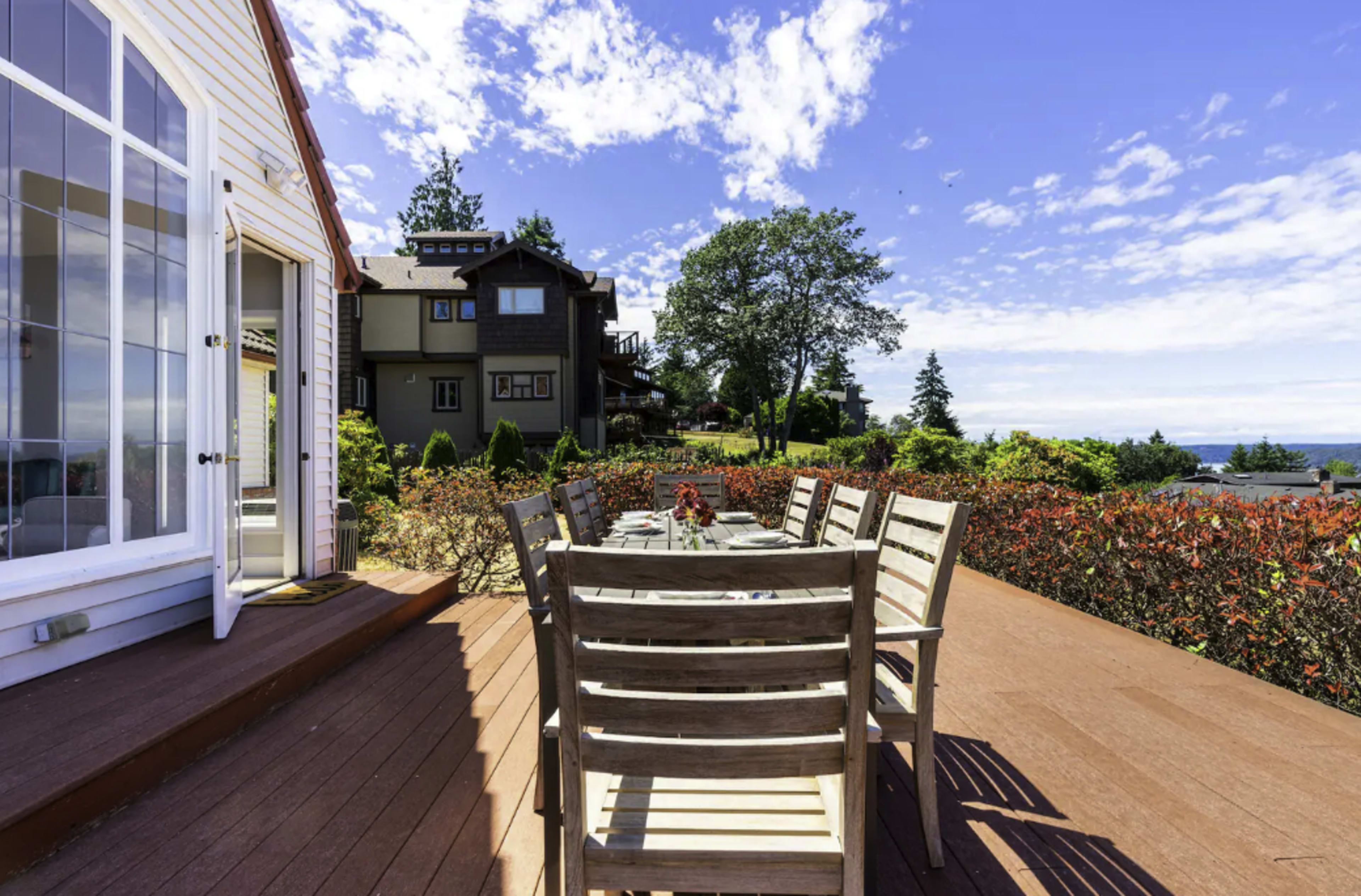A wooden deck with a large dining table and chairs overlooks a landscaped garden and houses under a clear blue sky.