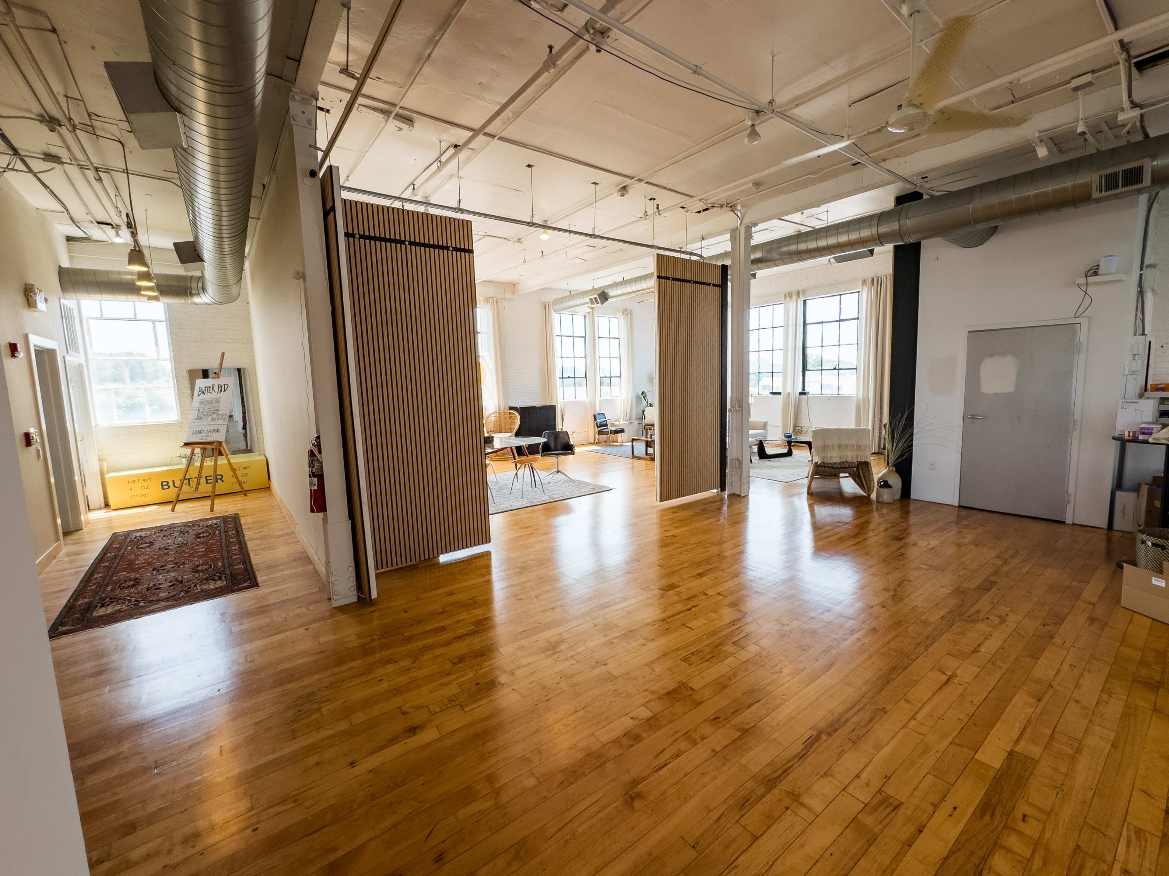 The image shows a spacious, modern interior with wooden floors, large windows, and multiple seating areas separated by partition walls.