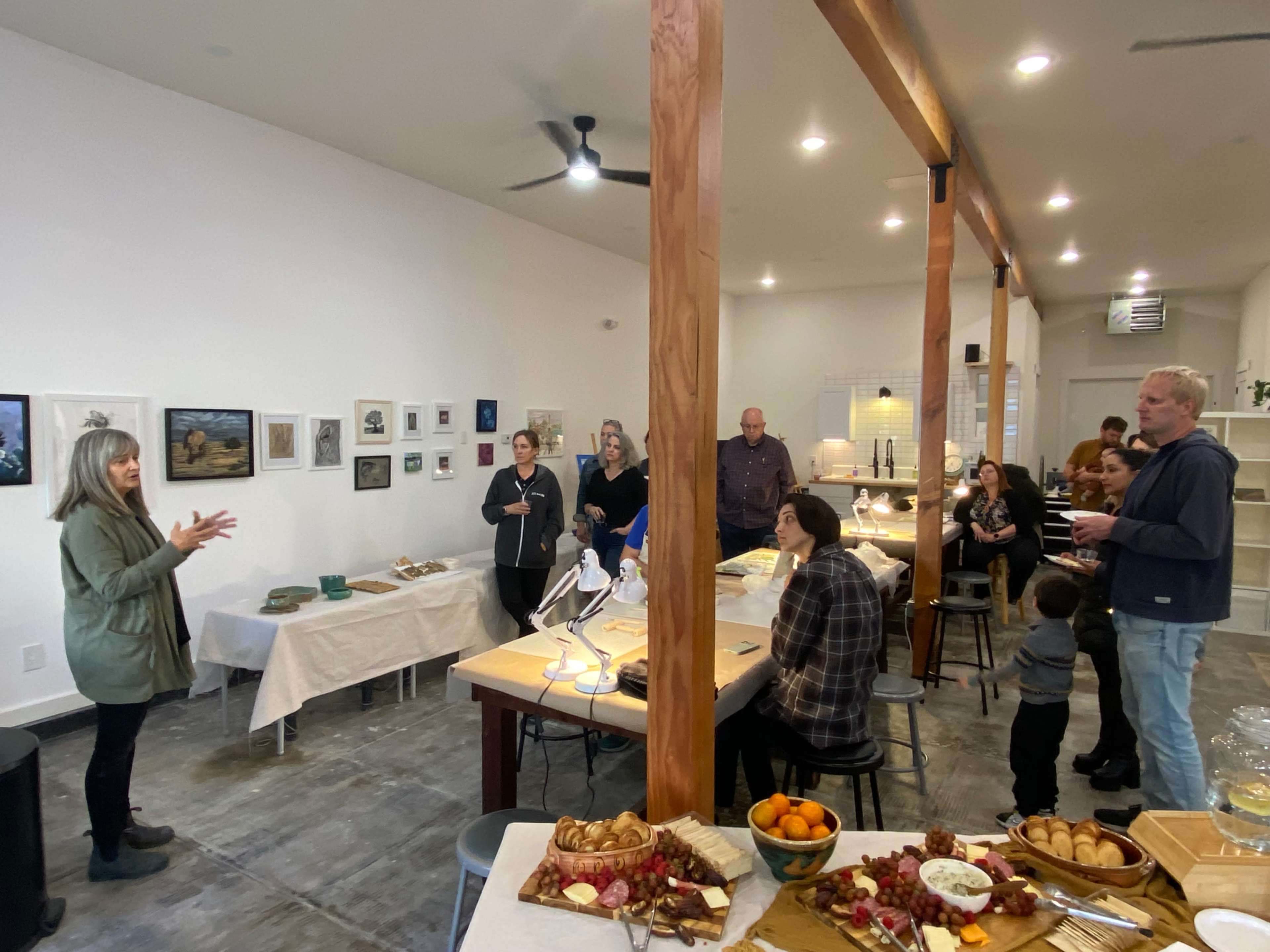A woman stands and speaks to a small group in a modern gathering space with artwork on the walls and a table laden with food.