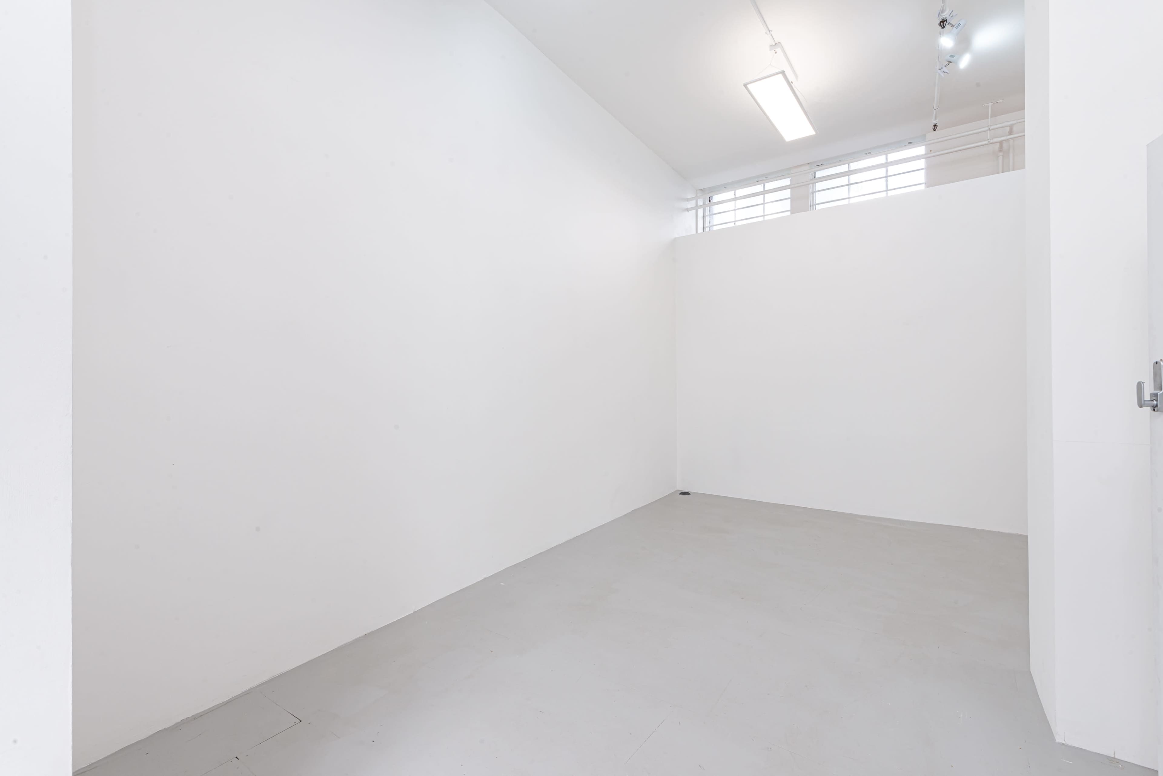 An empty white room with bare walls and a gray floor, illuminated by overhead lights.