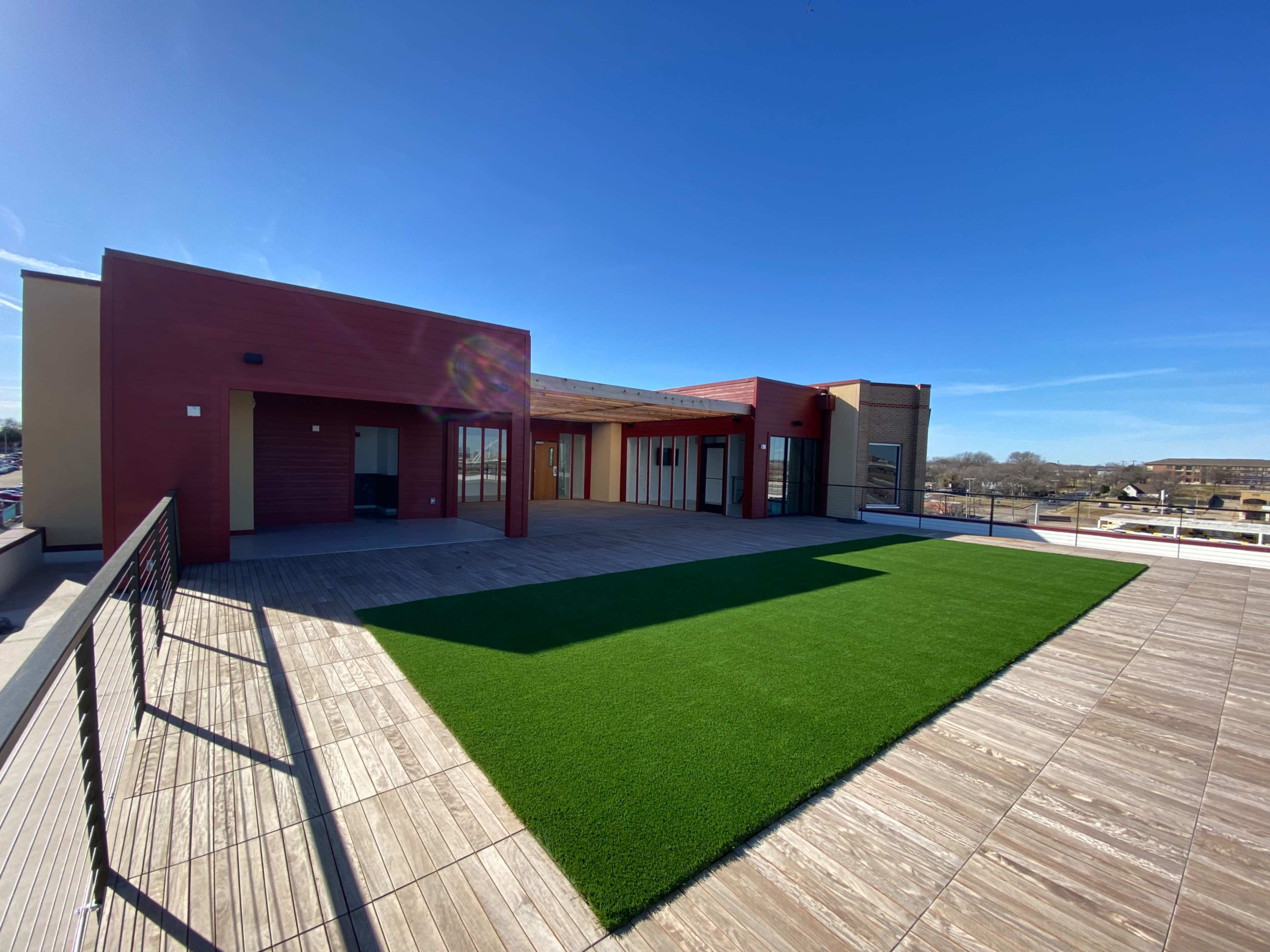 The image shows a rooftop space with a green artificial grass area and wooden decking, alongside modern buildings under a clear blue sky.