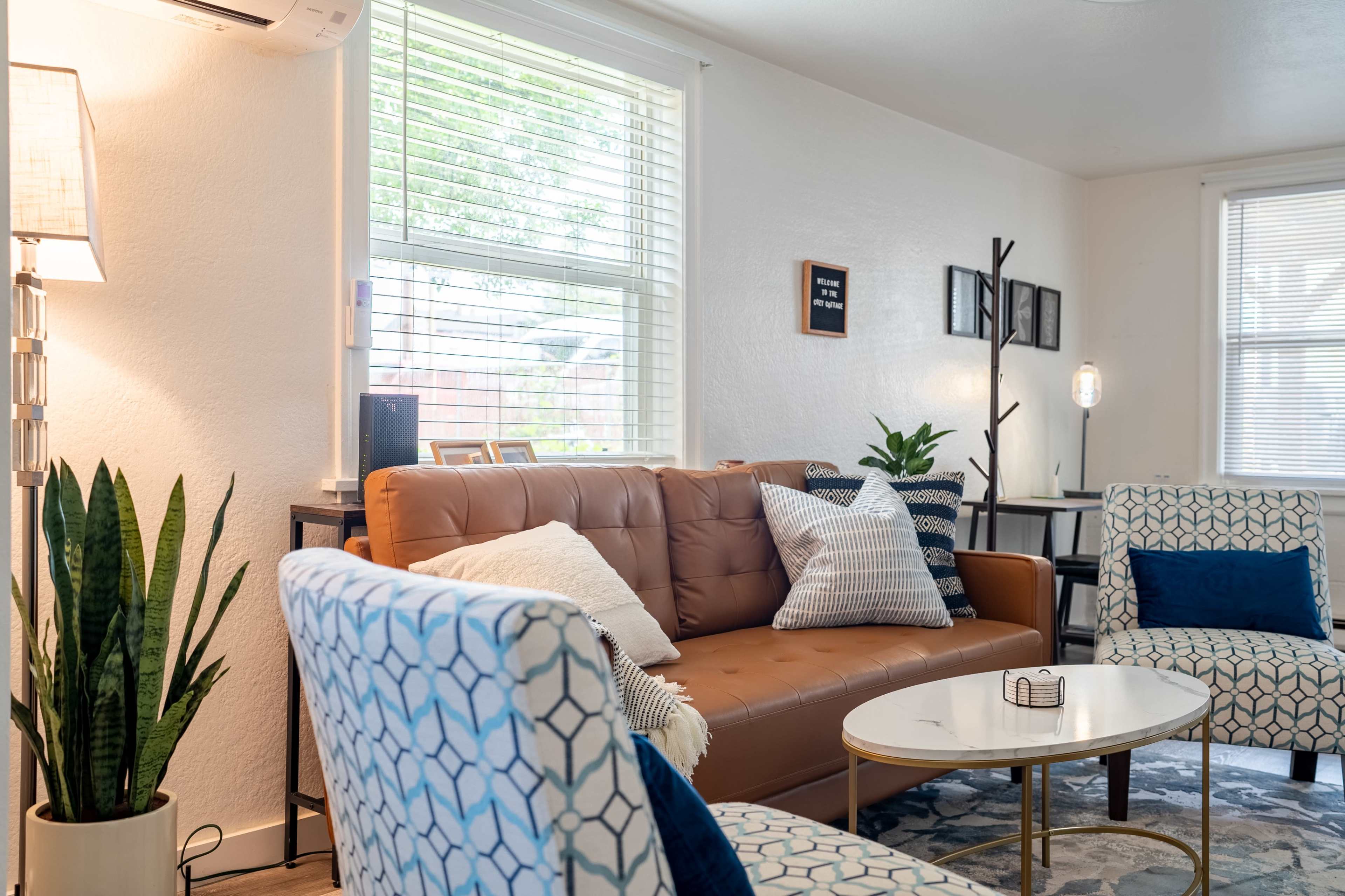 A bright living room with a brown sofa, two patterned chairs, a round coffee table, and a large window allowing natural light to enter.