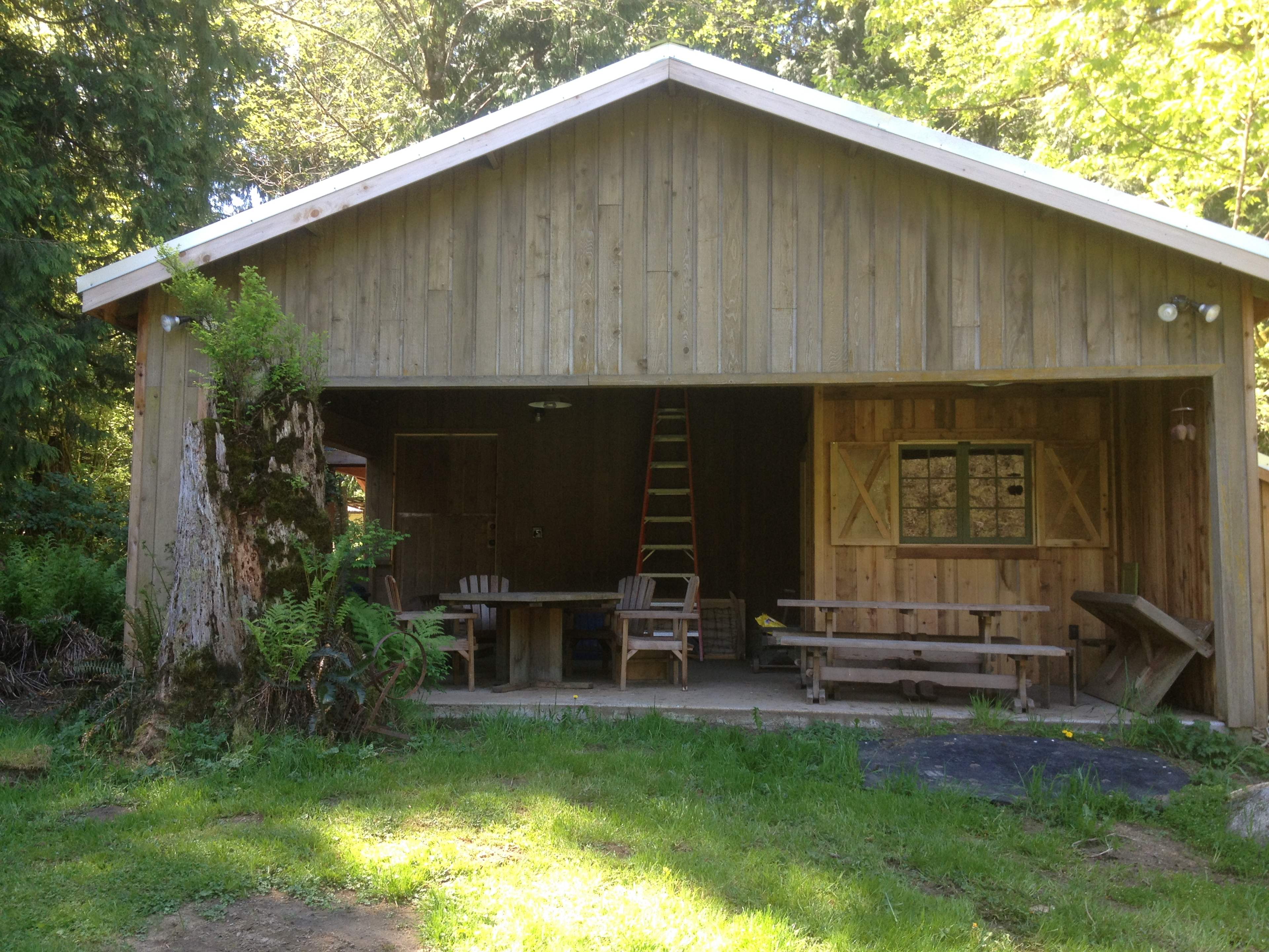 A wooden cabin with a covered porch, surrounded by greenery and featuring picnic tables and a ladder against the wall.