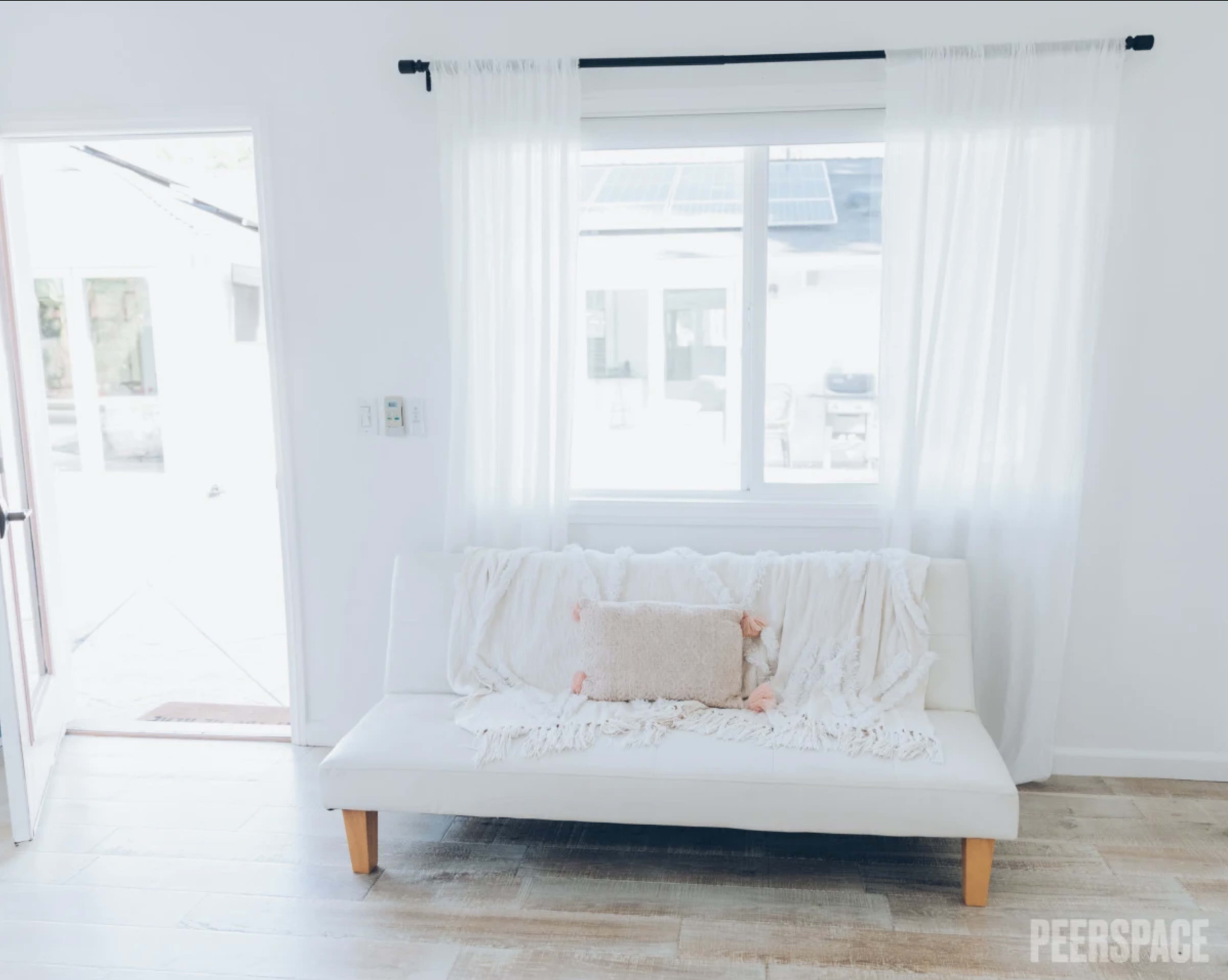 A white couch with a light blanket and decorative pillow is positioned in front of a large window with sheer curtains, illuminating a bright, minimalist room.