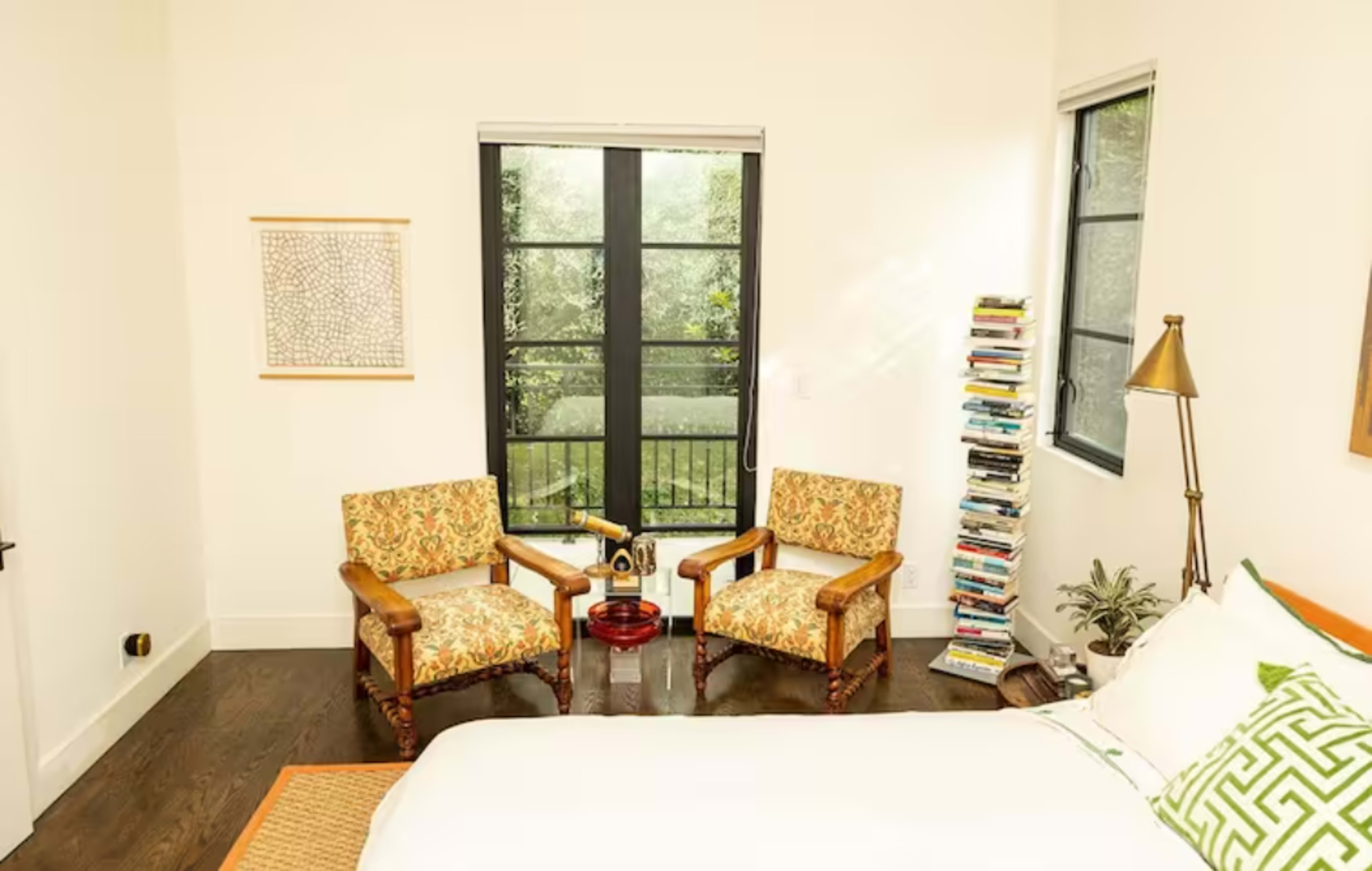The room features a bed, two patterned armchairs, a stack of books positioned by the window, and greenery visible outside.