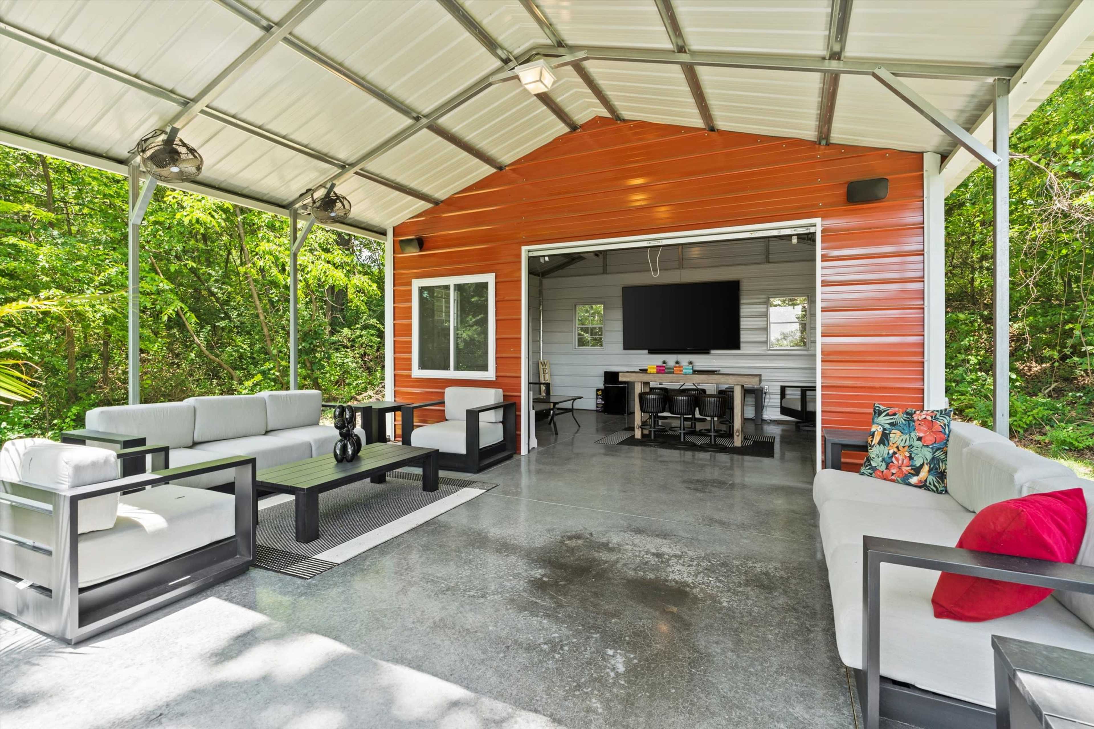 The image shows a covered outdoor seating area with a television, a dining table, and several modern sofas arranged on a concrete floor surrounded by greenery.