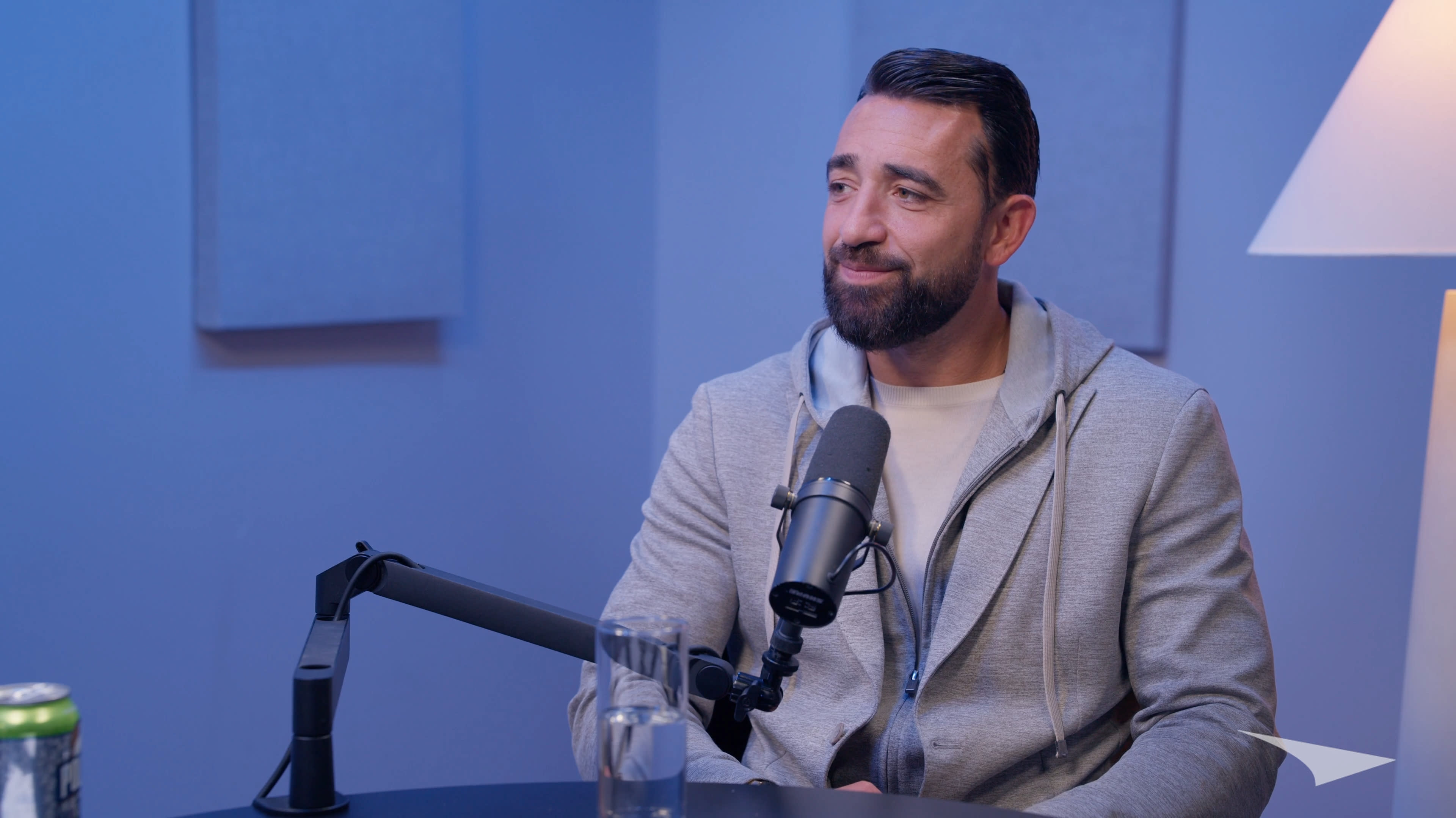 A man with a beard is sitting at a table with a microphone in front of him and a glass of water nearby.