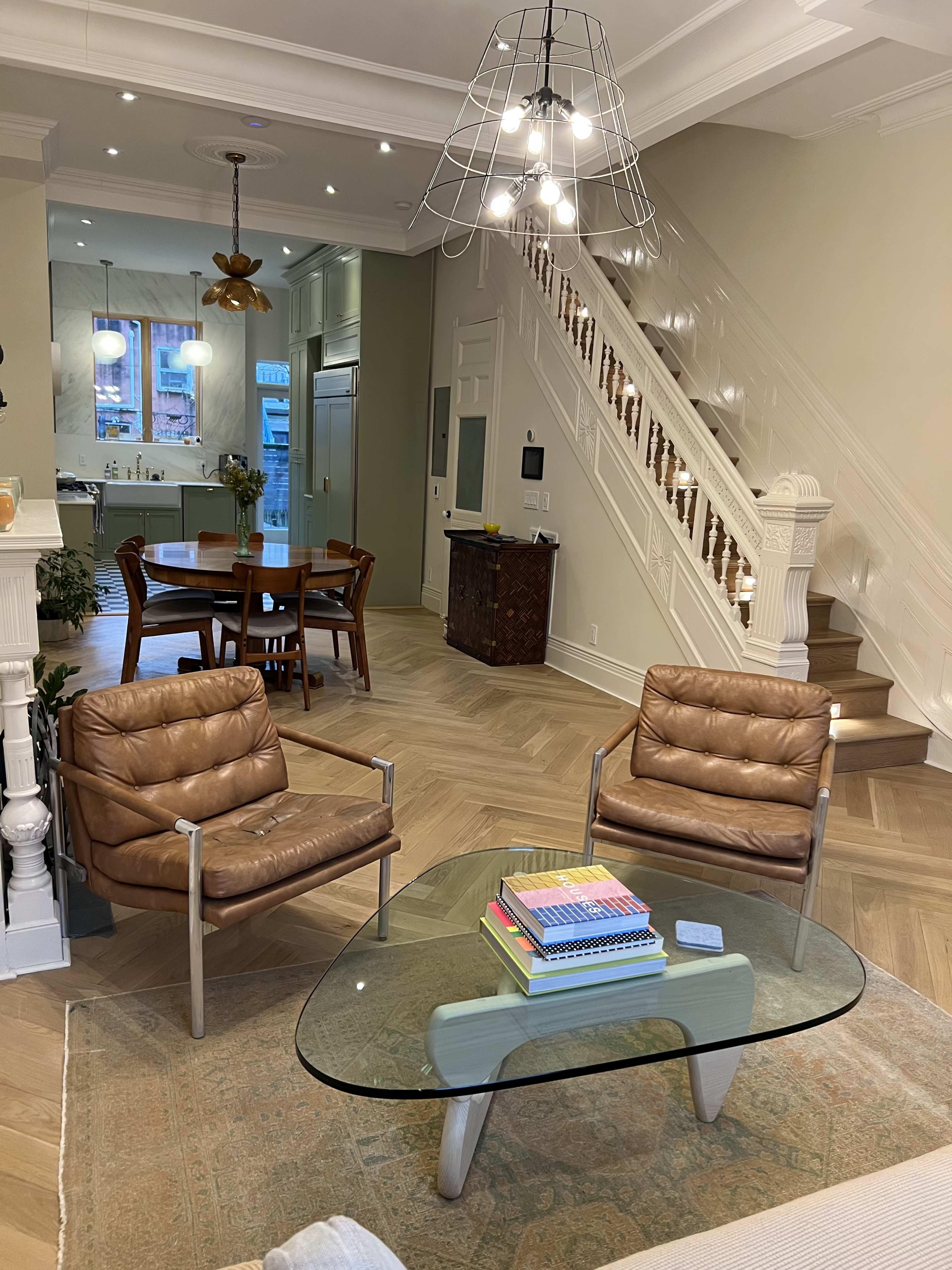 A modern living room featuring two brown leather chairs, a glass coffee table with books, and a visible dining area and staircase in the background.