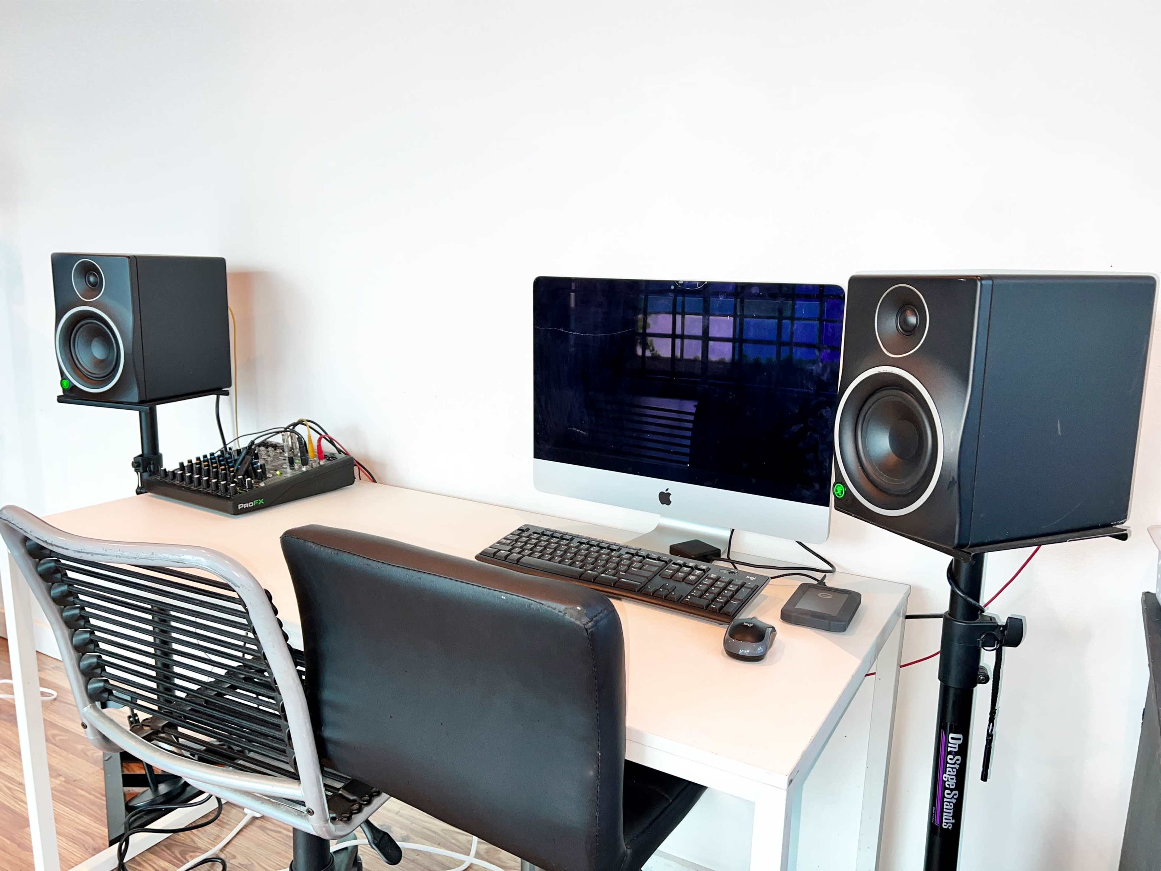 A desk with an iMac, a keyboard, a mouse, and two studio speakers is set up against a white wall.