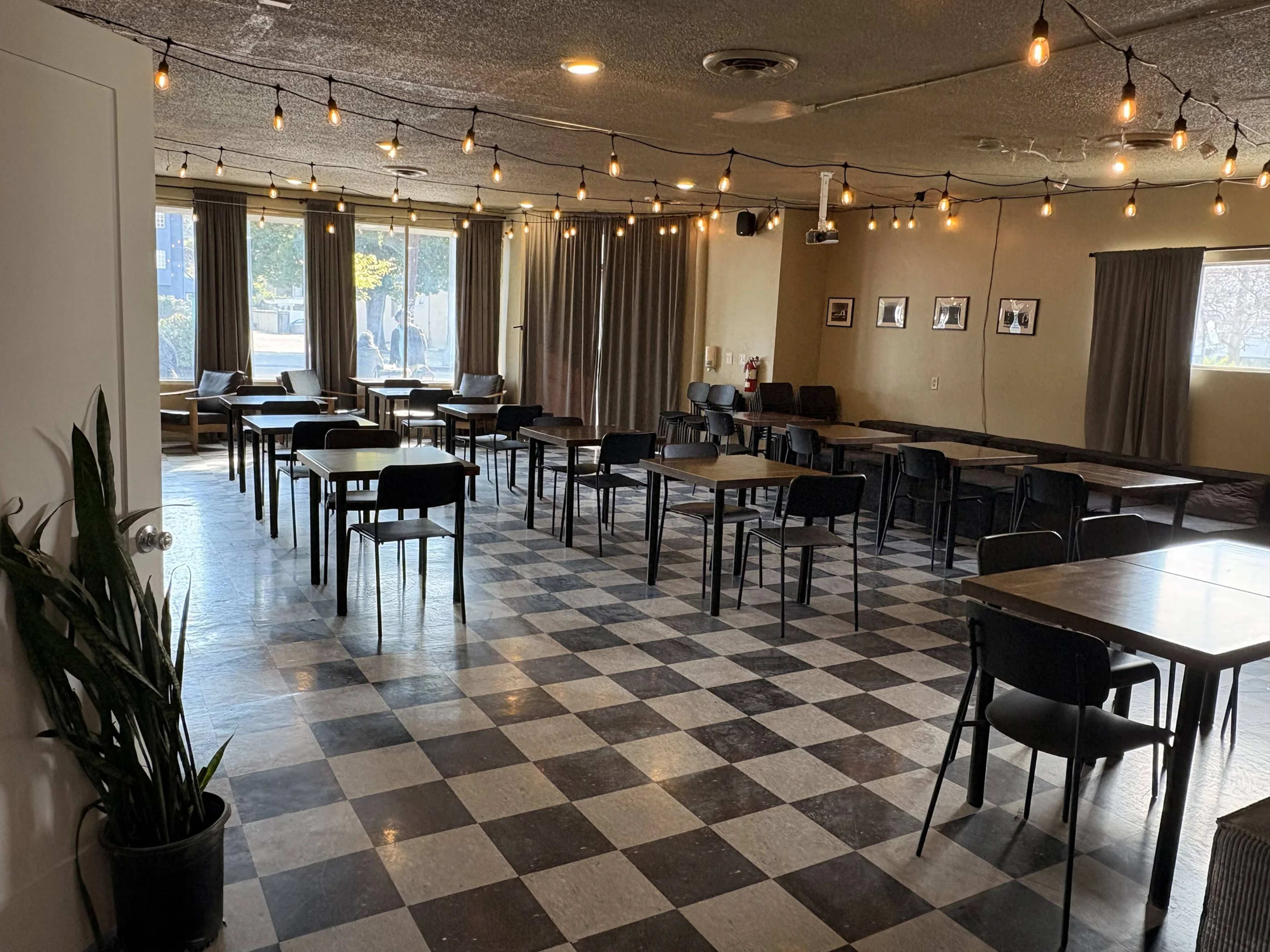 The image shows a spacious dining area with a checkered floor, several tables and chairs arranged in rows, and string lights hanging from the ceiling.