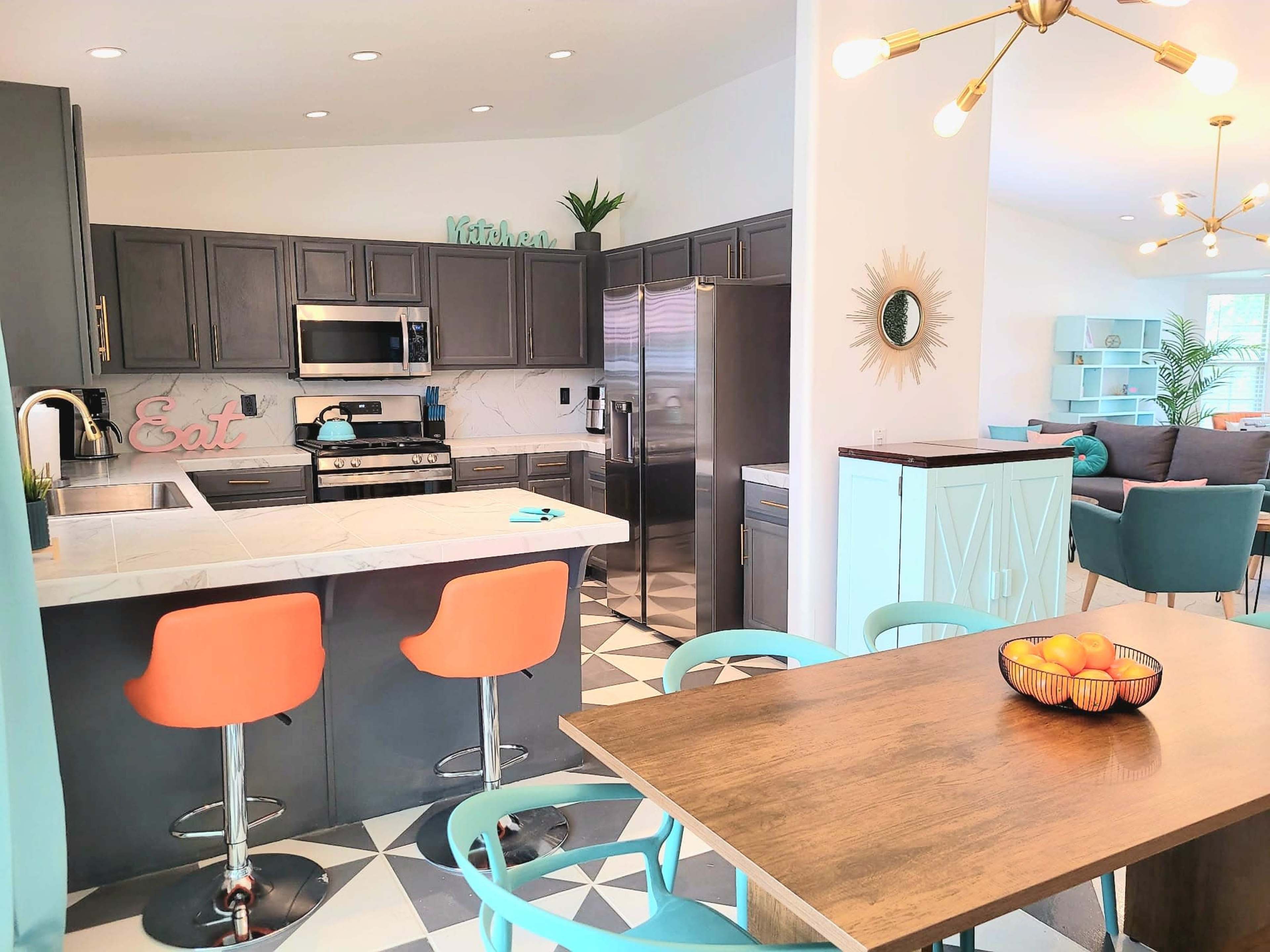 A modern kitchen with gray cabinets, stainless steel appliances, and a dining area featuring a wooden table and colorful bar stools.