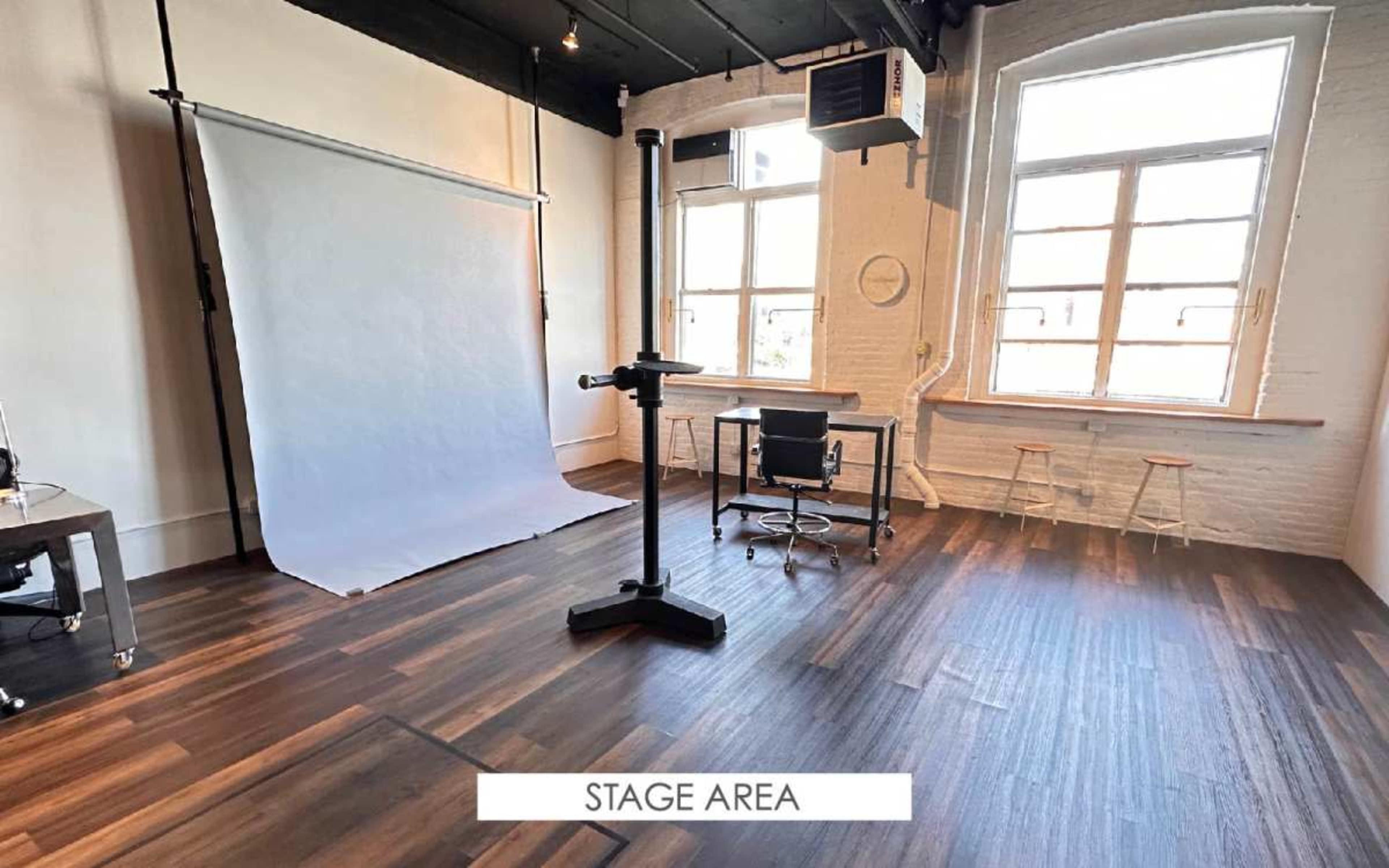 The image shows a spacious, well-lit studio with wooden flooring, featuring a blank backdrop, a rolling stand, a chair, and a small table.