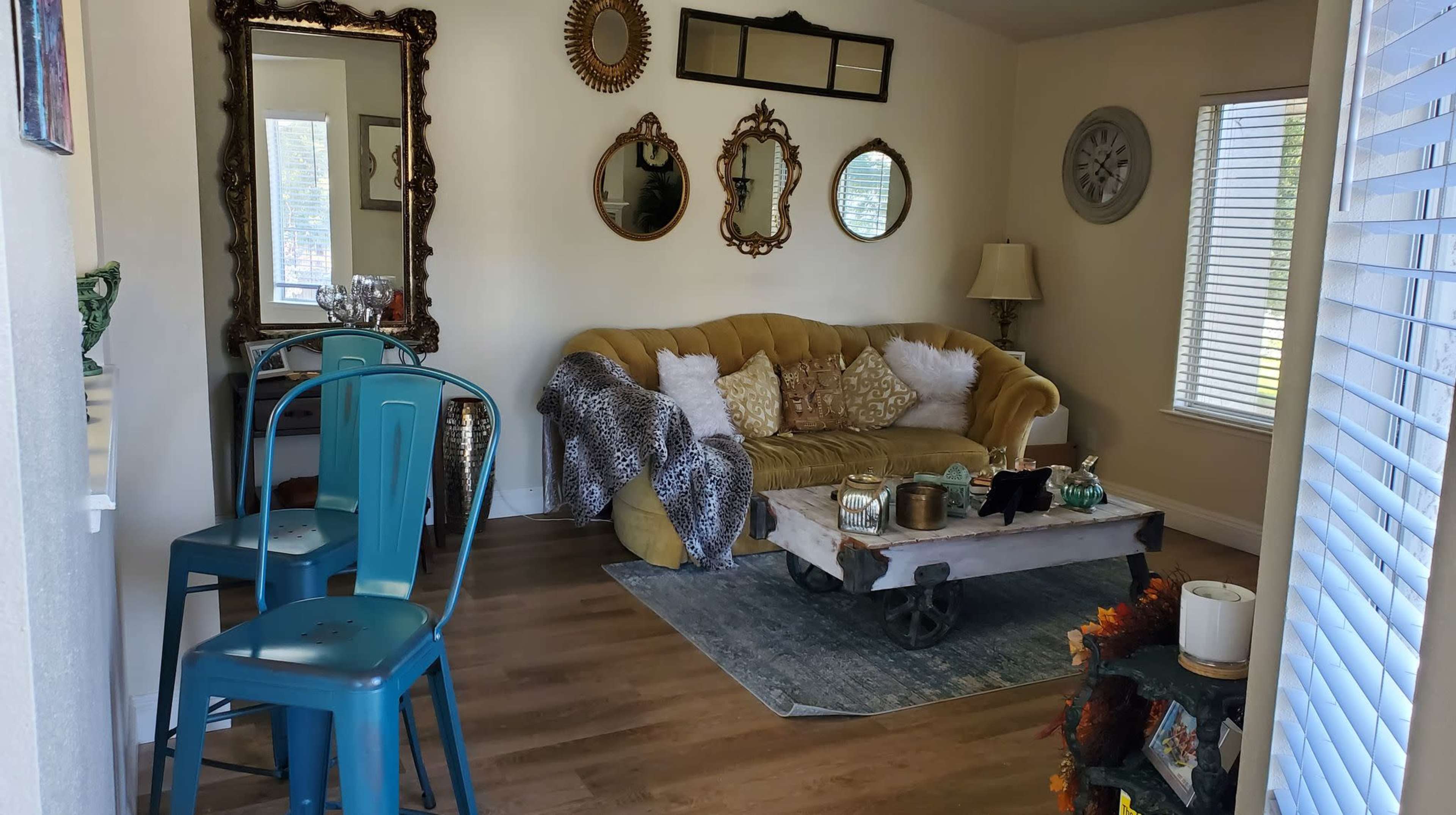 A cozy living room featuring a yellow couch with a patterned throw, a rustic coffee table on a blue rug, and several wall mirrors.
