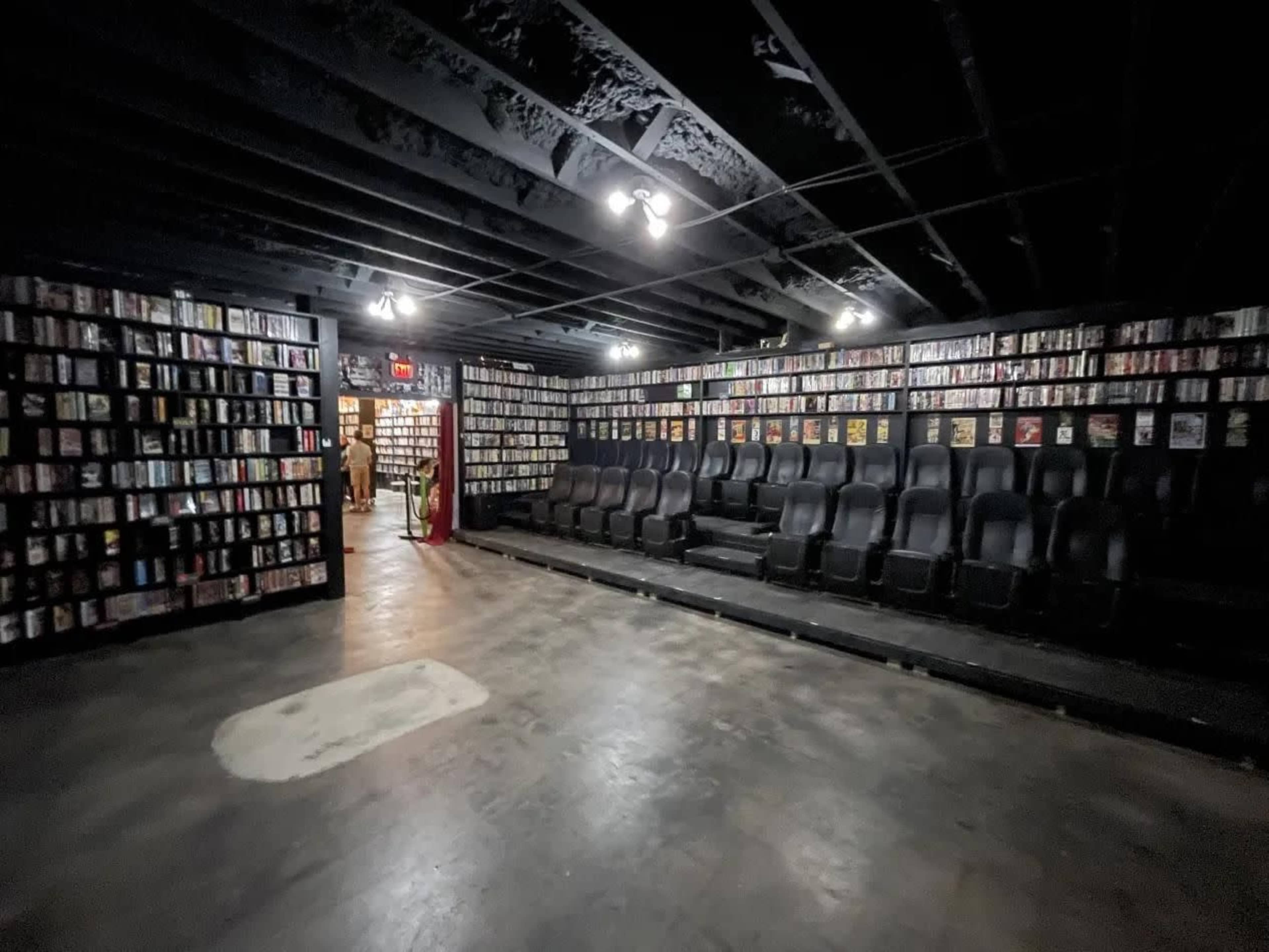 The dimly lit space features shelves lined with DVDs and a row of black theater seats facing them.