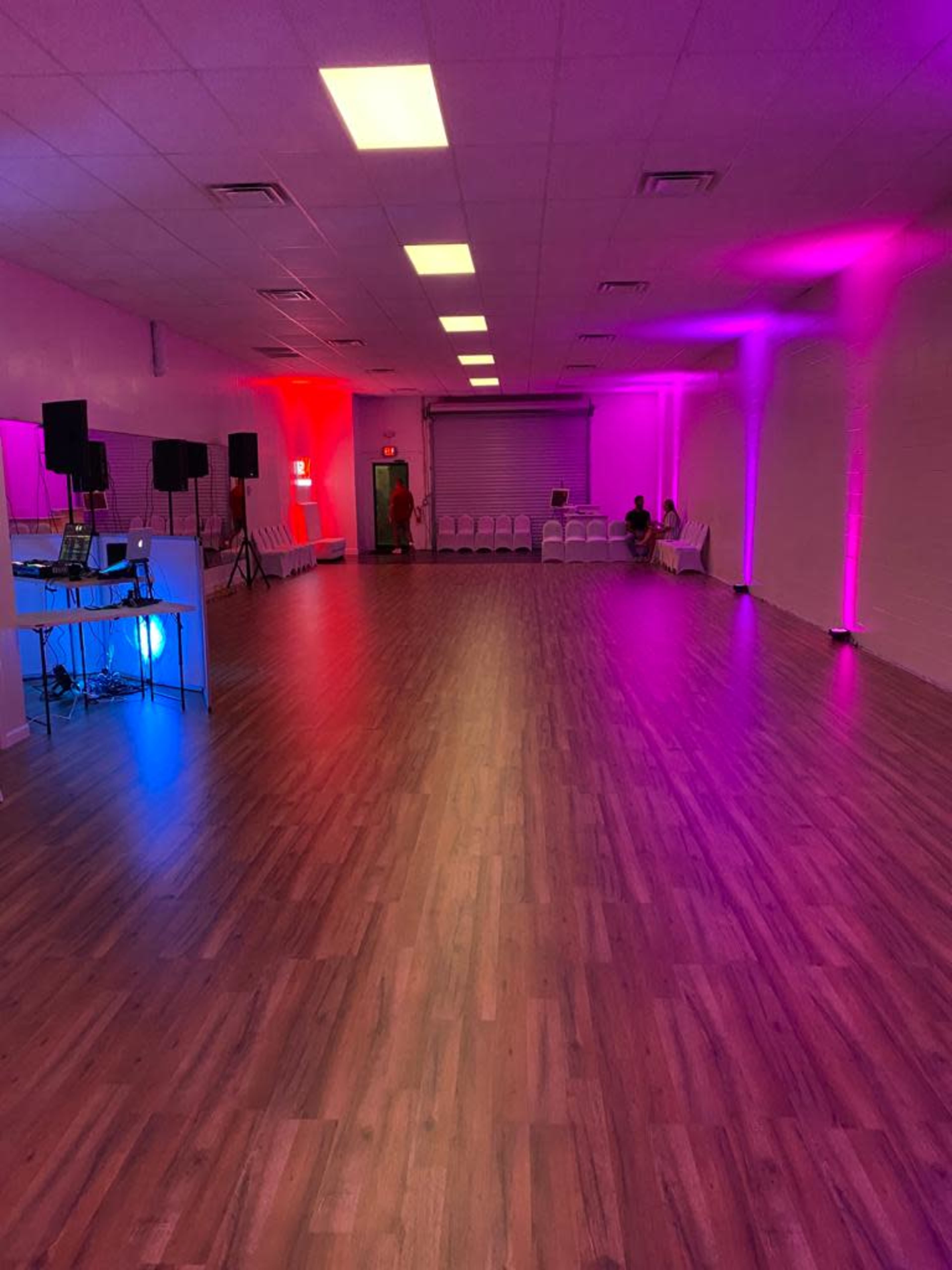 A spacious room with a polished wooden floor, featuring colorful lighting along the walls and a setup for music equipment at one end.