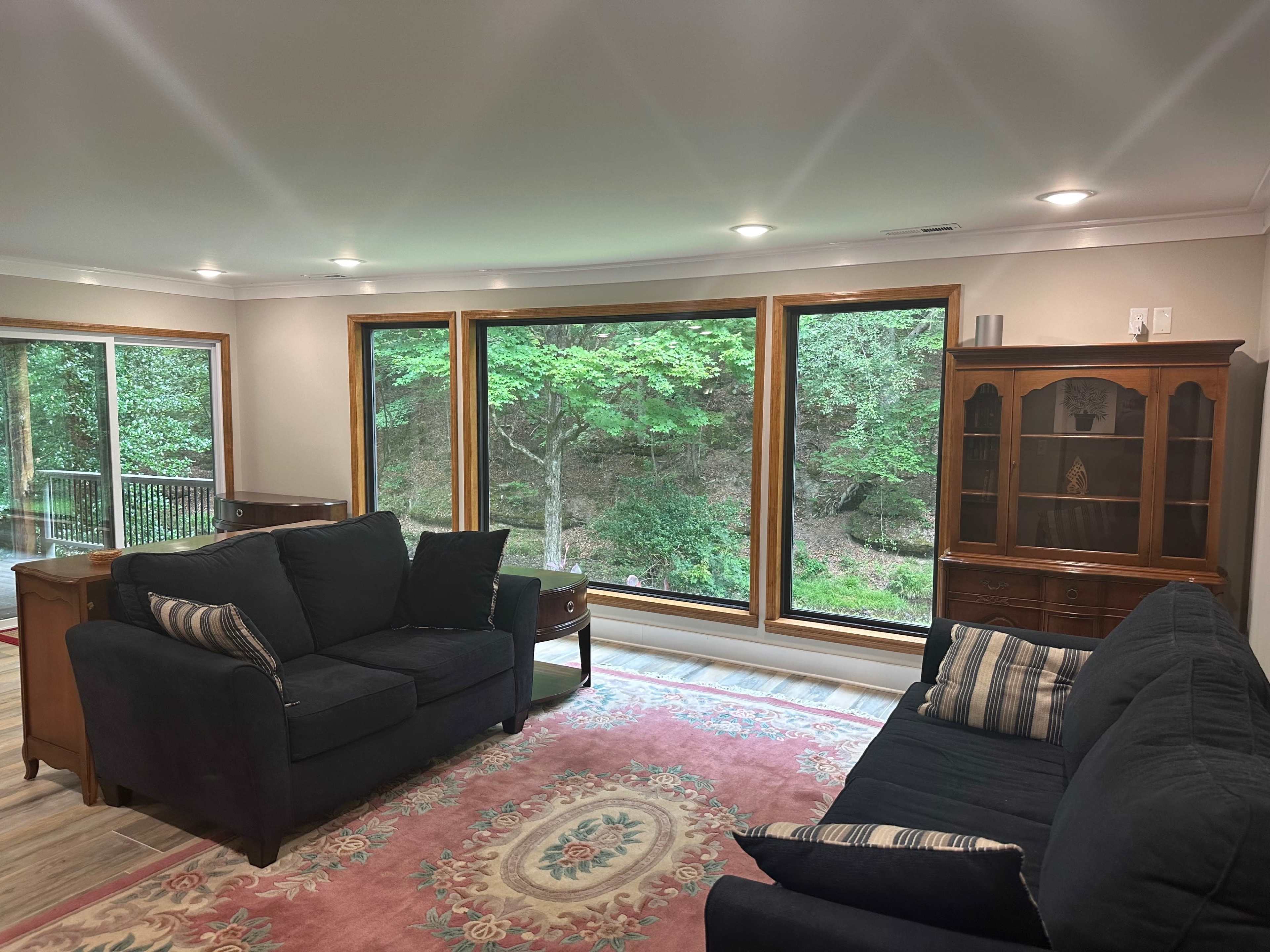 The living room features two dark sofas facing large windows that overlook a wooded area, with a rug and a wooden cabinet in the space.