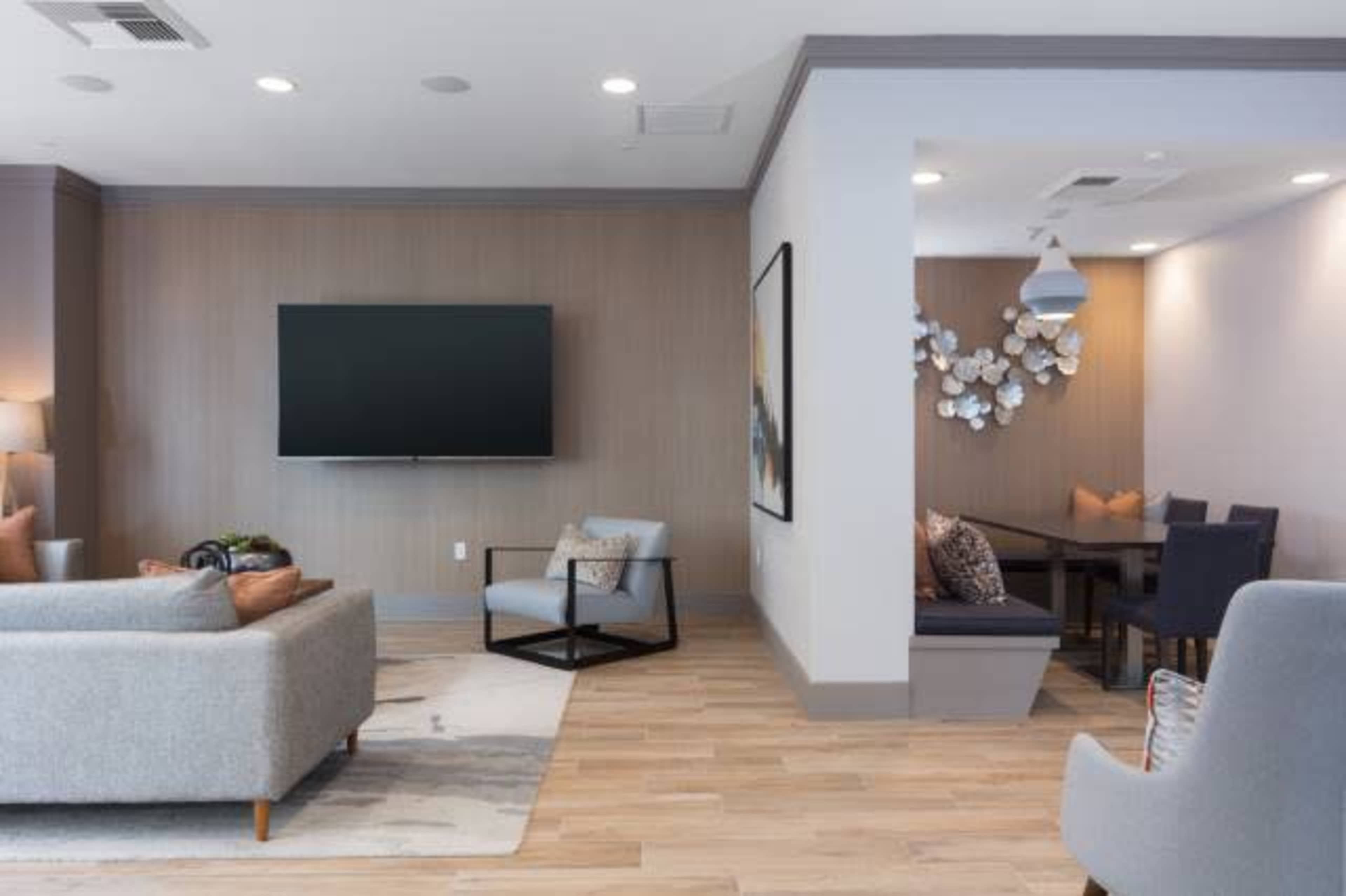 A modern living area with a flat-screen TV mounted on the wall, a gray sofa, a light wood floor, and a dining area with a table and seating.